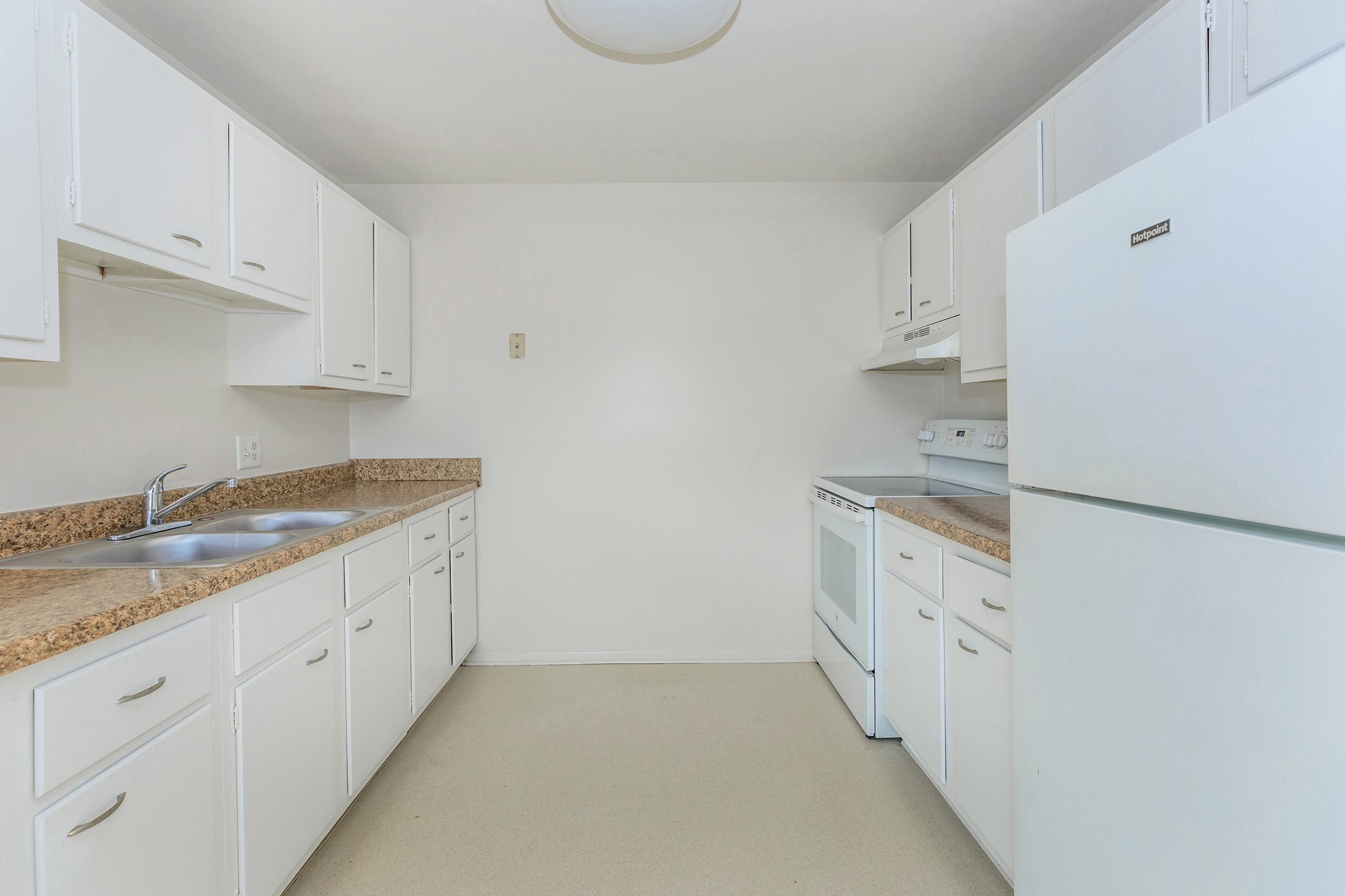 A clean, modern kitchen featuring white cabinets, a double sink, a white stove, and a refrigerator. The countertops are a light-colored stone, and there is a neutral-colored floor. The walls are painted white, creating a bright and spacious atmosphere.