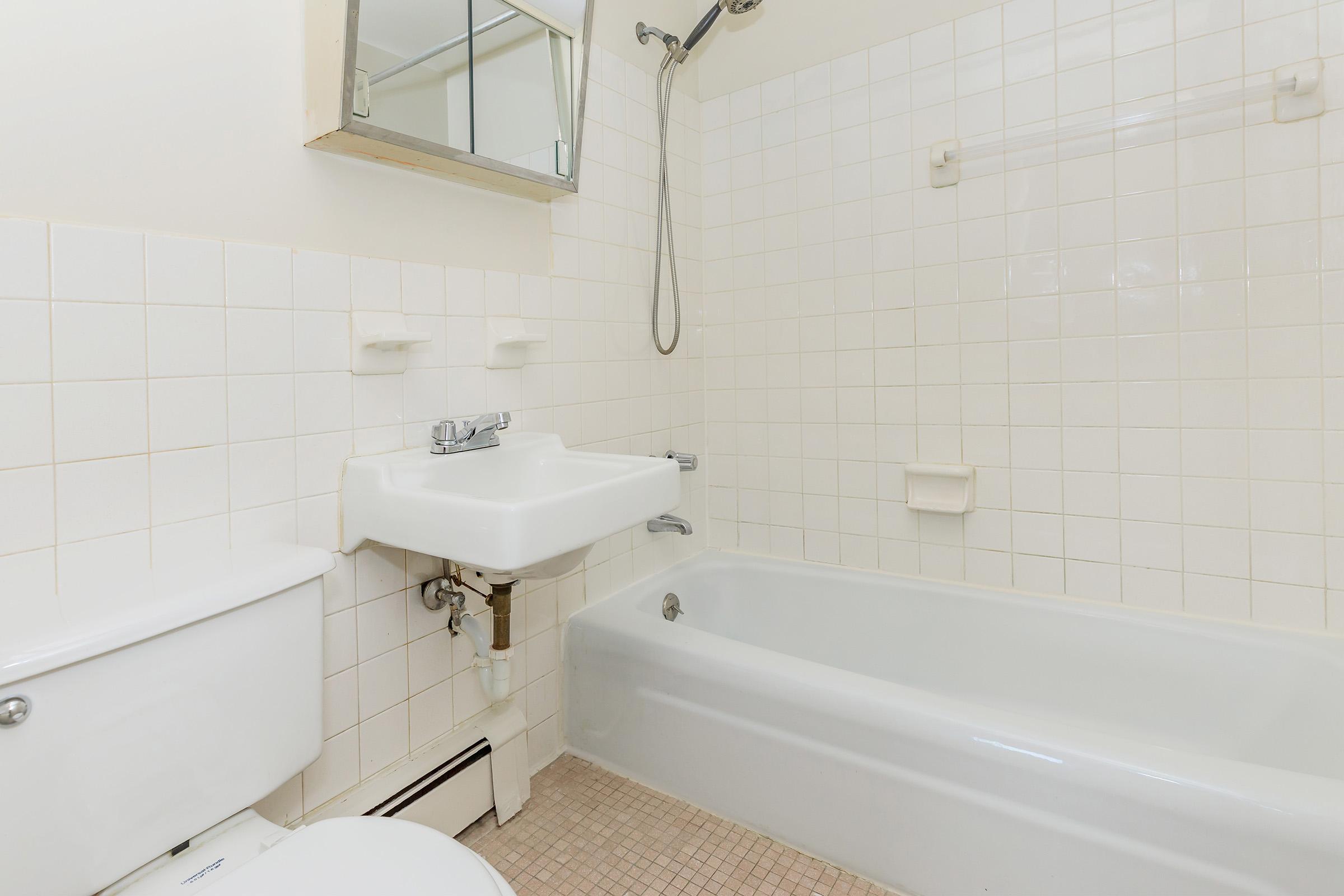 A clean and simple bathroom featuring a white bathtub, a wall-mounted shower with a showerhead, a sink with a mirror above it, and a toilet. The walls are tiled with light-colored square tiles and the floor is covered in small beige tiles. Natural light illuminates the space.