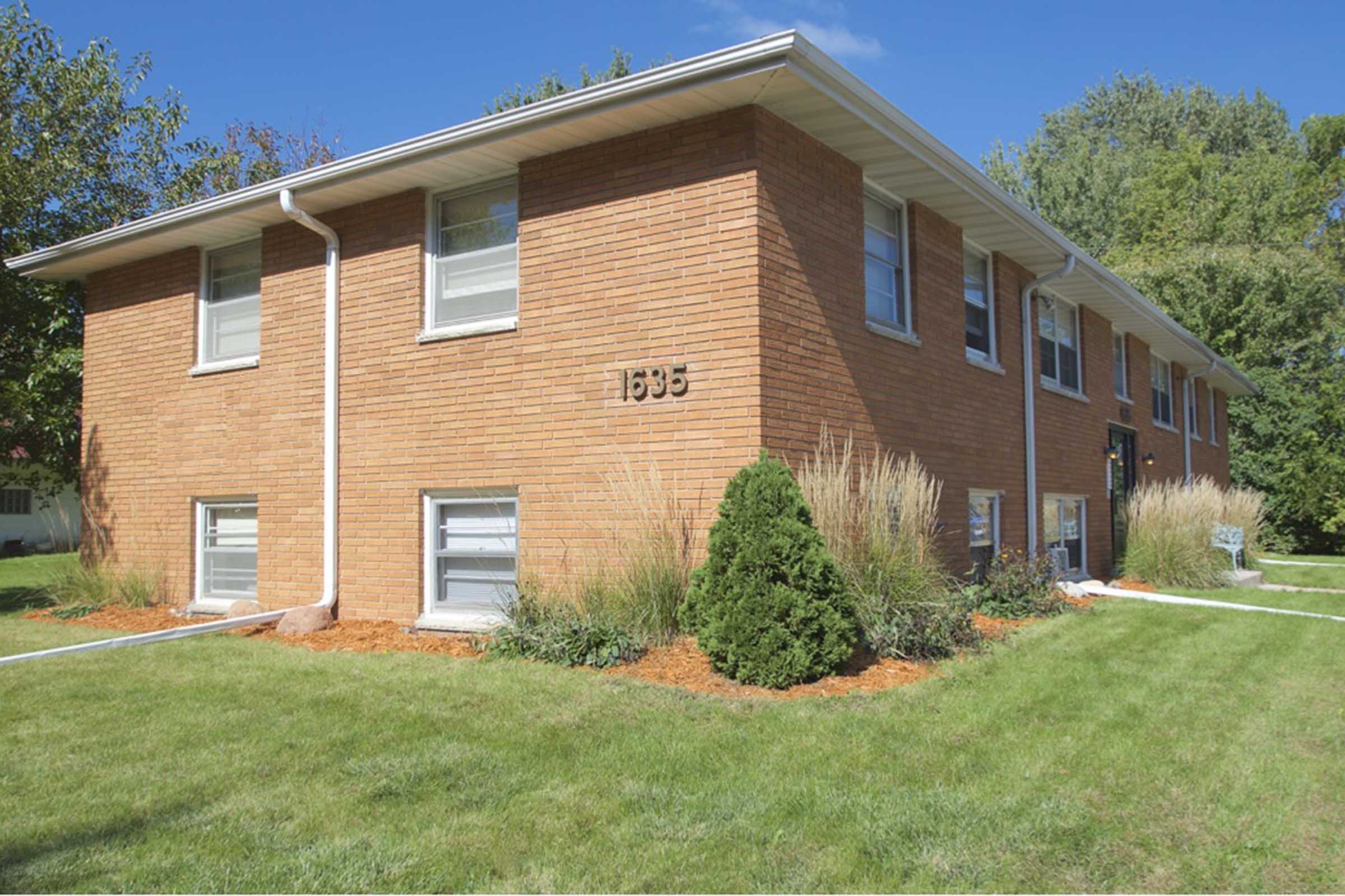 A brick apartment building featuring multiple windows and a neatly landscaped front yard with grass and shrubs. The building has a homey atmosphere, with trees in the background under a clear blue sky.