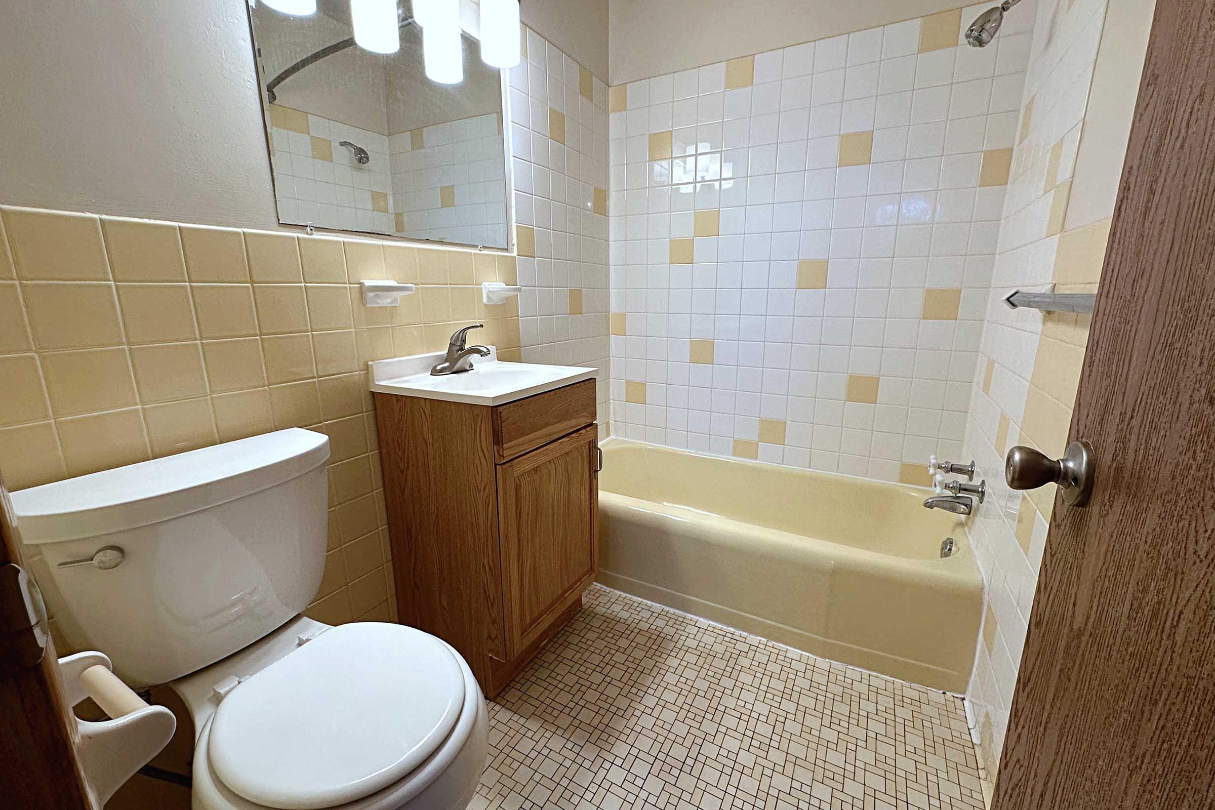 A small bathroom featuring a yellow and white tiled design. It includes a bathtub with a shower above, a wooden vanity with a sink, a toilet, and a mirror above the sink. The floor is covered in small tiles, contributing to a vintage aesthetic. The lighting is provided by a ceiling light fixture.