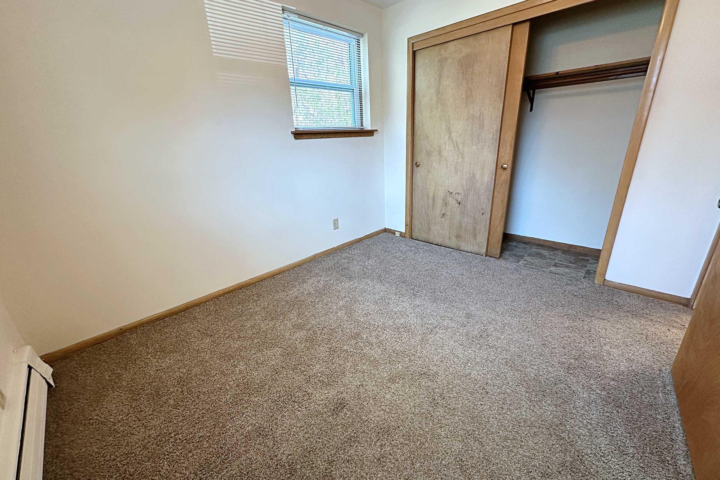 Empty room with beige carpet and white walls. A window on the left side allows natural light in. There is a wooden closet on the right with doors closed and space for hanging clothes. The overall appearance is clean and minimalistic, suitable for a bedroom or office space.