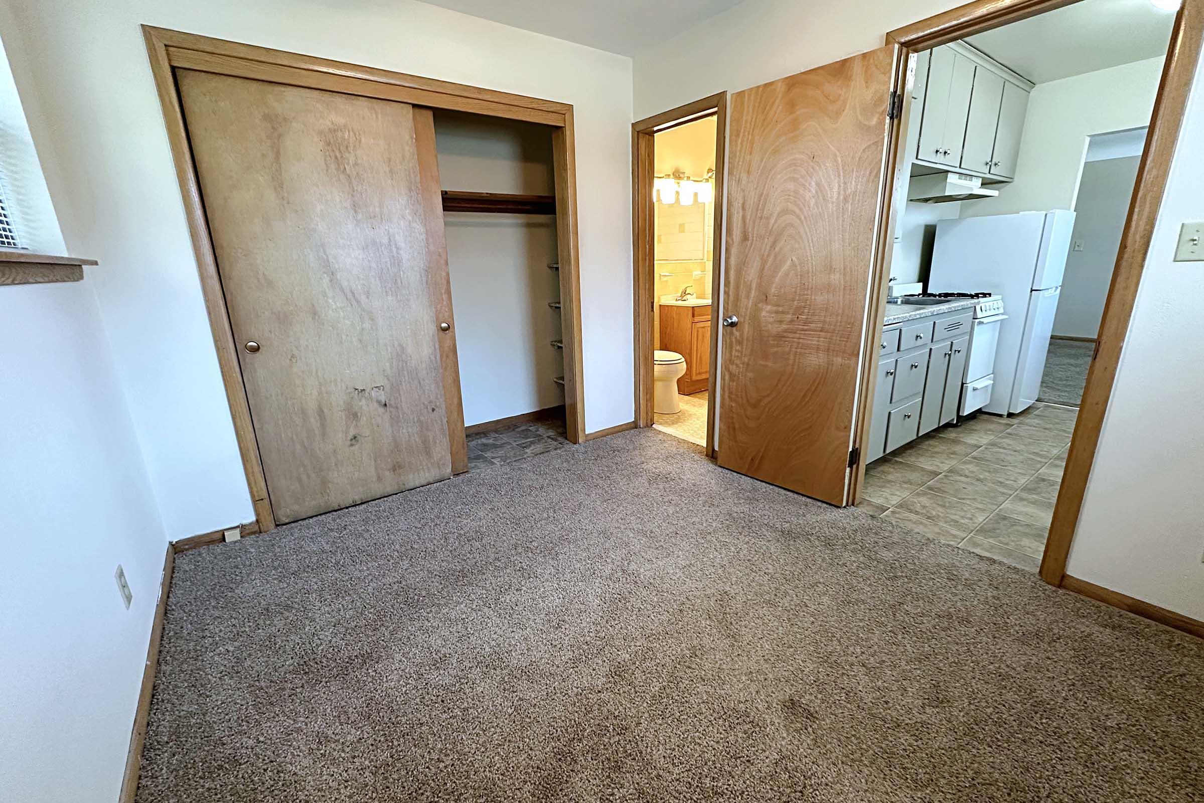 A cozy room with beige carpet, featuring two wooden doors—one leading to a closet and the other to a hallway. In the background, a glimpse of a bathroom can be seen through an open door. The room has neutral-colored walls and ample natural light.