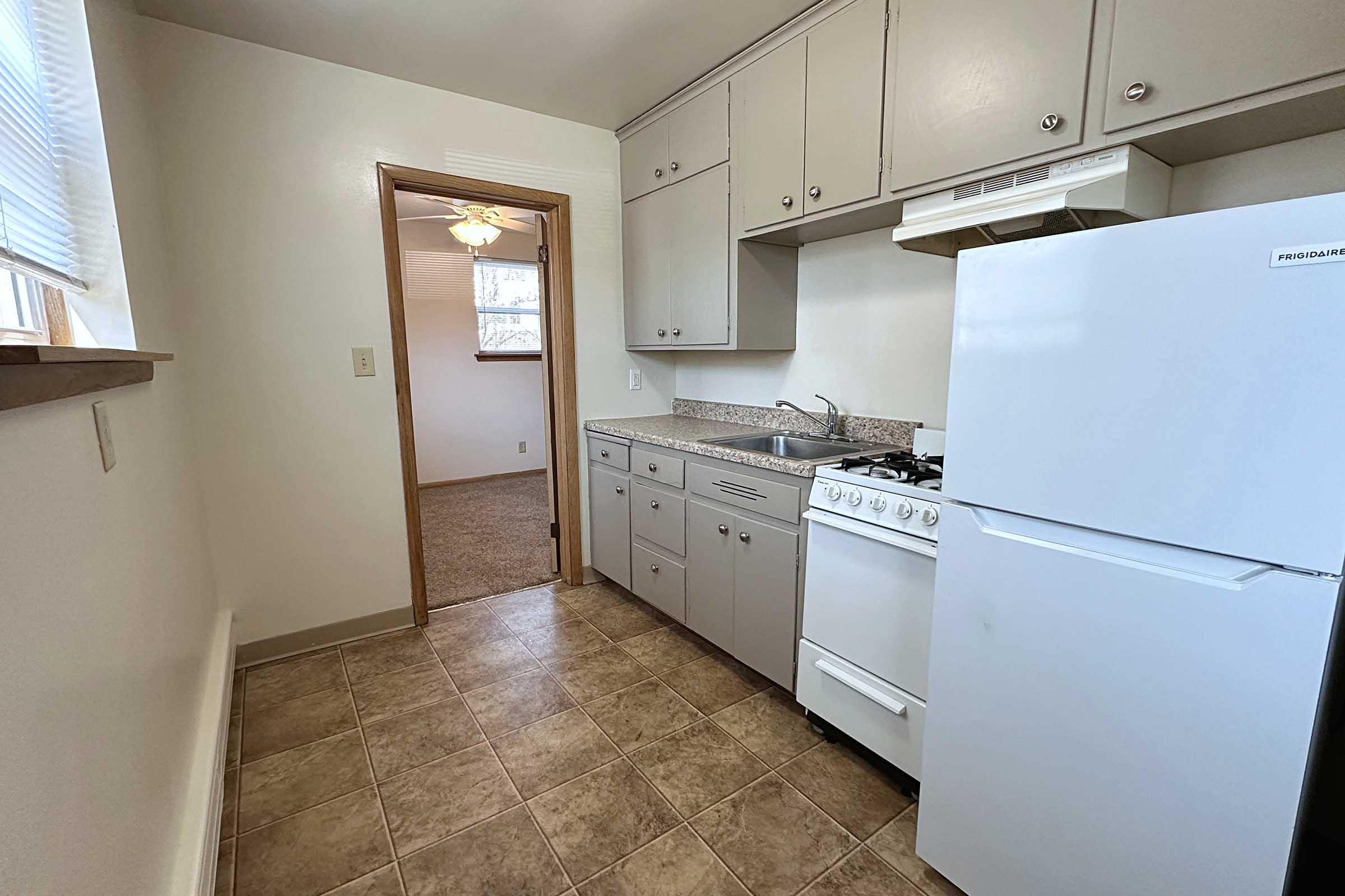 A small kitchen featuring light-colored cabinets, a white refrigerator, a gas stove, and a countertop with a sink. The floor is tiled, and there's a door leading to another room, with a ceiling fan visible in the background. Natural light comes in through a window.