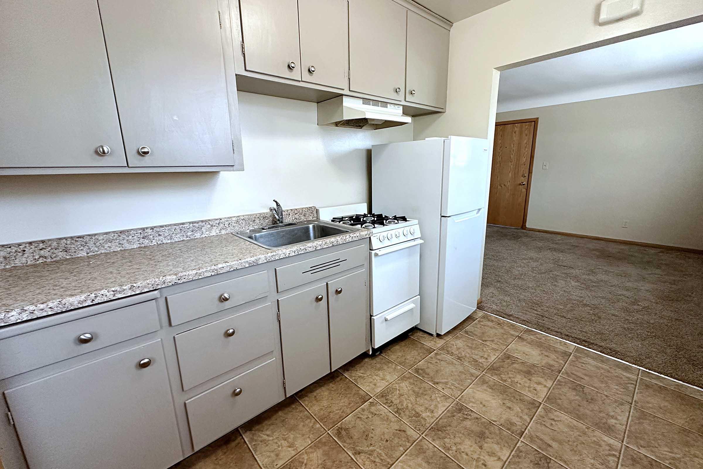 A compact kitchen featuring light gray cabinets, a countertop with a sink, a gas stove, and a white refrigerator. The kitchen opens to a carpeted living area in the background, with a wooden door visible. The overall design is simple and functional.