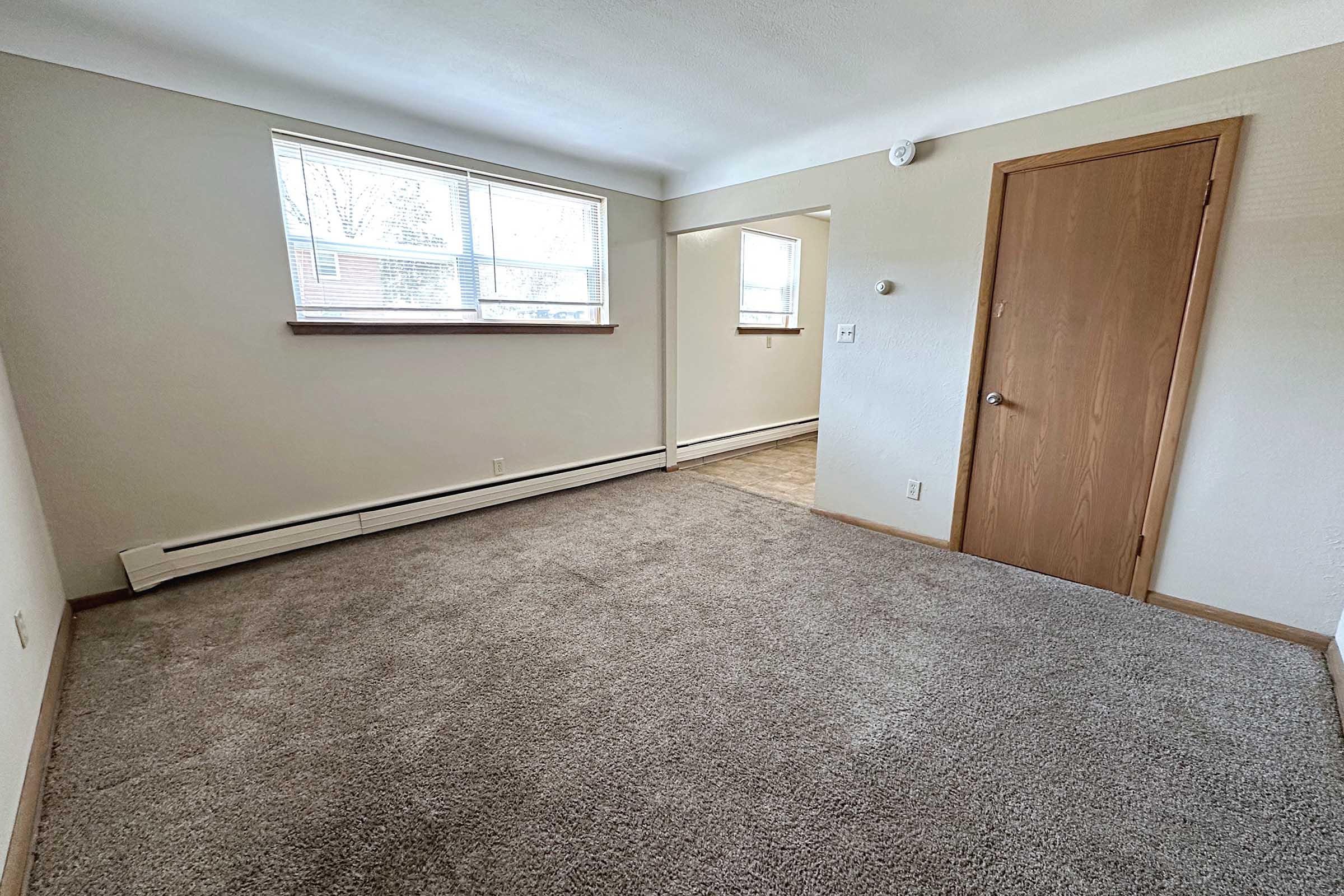 A well-lit, empty living room with light beige walls and a carpeted floor. It features a window with wooden blinds, revealing natural light. A doorway on the right leads to another room, and a baseboard heater is visible along the wall beneath the window. The space appears clean and ready for furnishing.