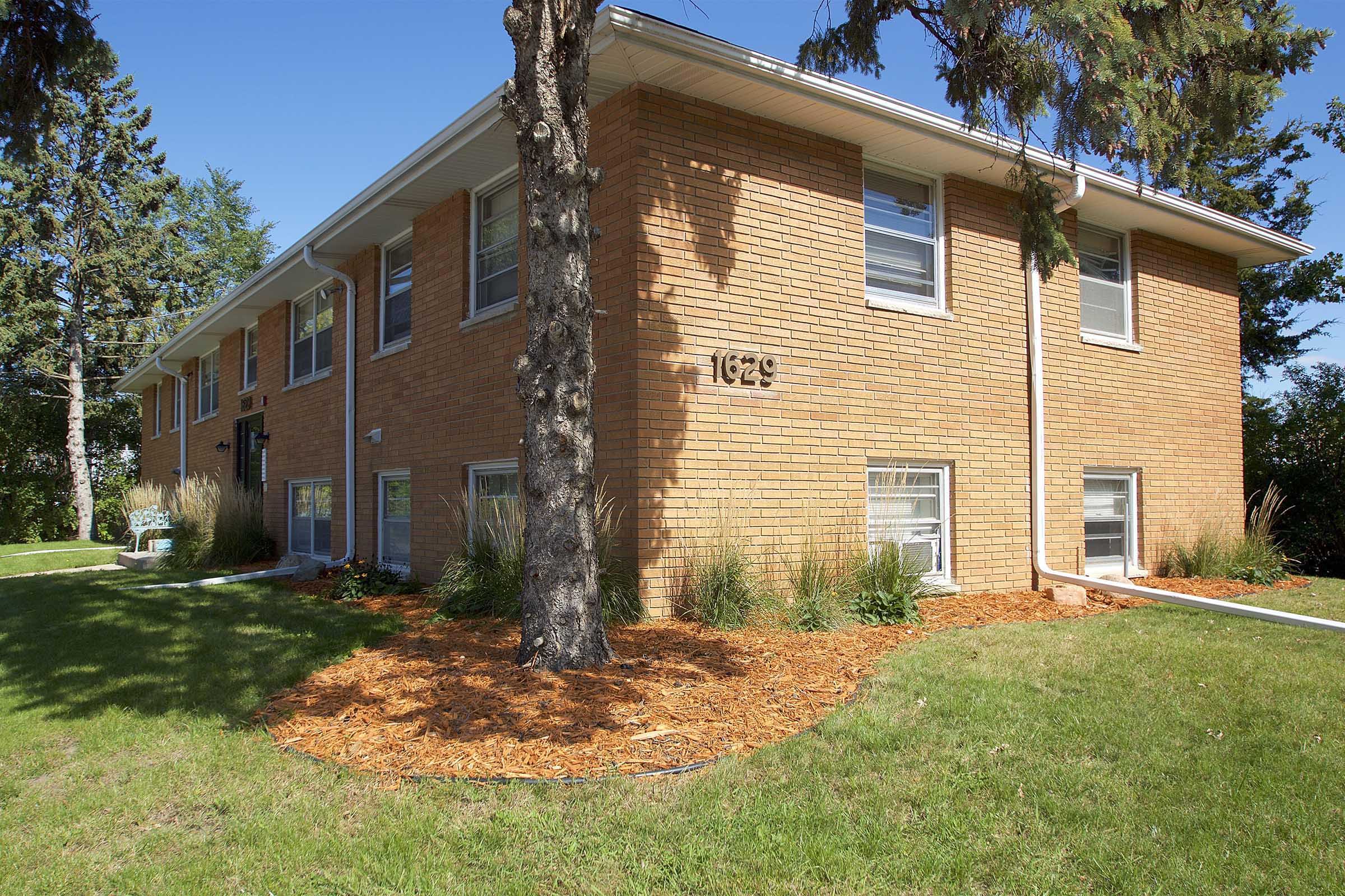 A two-story brick apartment building with a well-maintained lawn, bordered by trees and shrubs. The exterior features large windows and is adorned with fresh mulch around the base. The building number "1629" is visible on the front exterior. The sky is clear and blue.