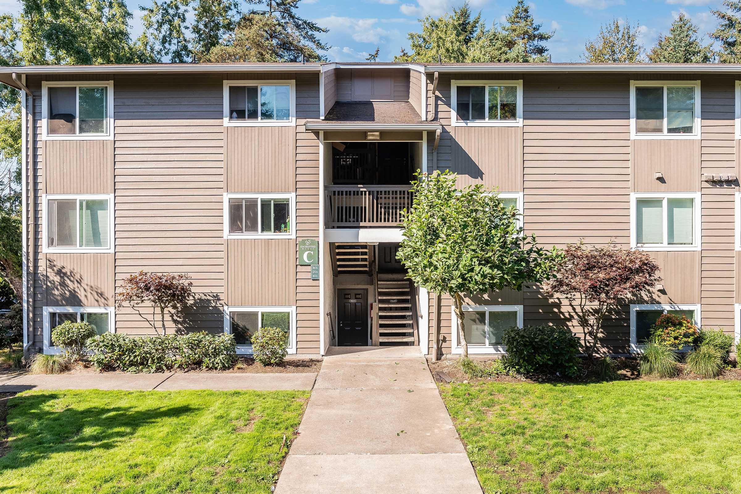 Three-story apartment building with light brown siding, featuring multiple windows and a central entrance with stairs leading to the upper floors. Green lawn and small shrubs frame the pathway leading to the front door. Trees and blue sky are visible in the background.