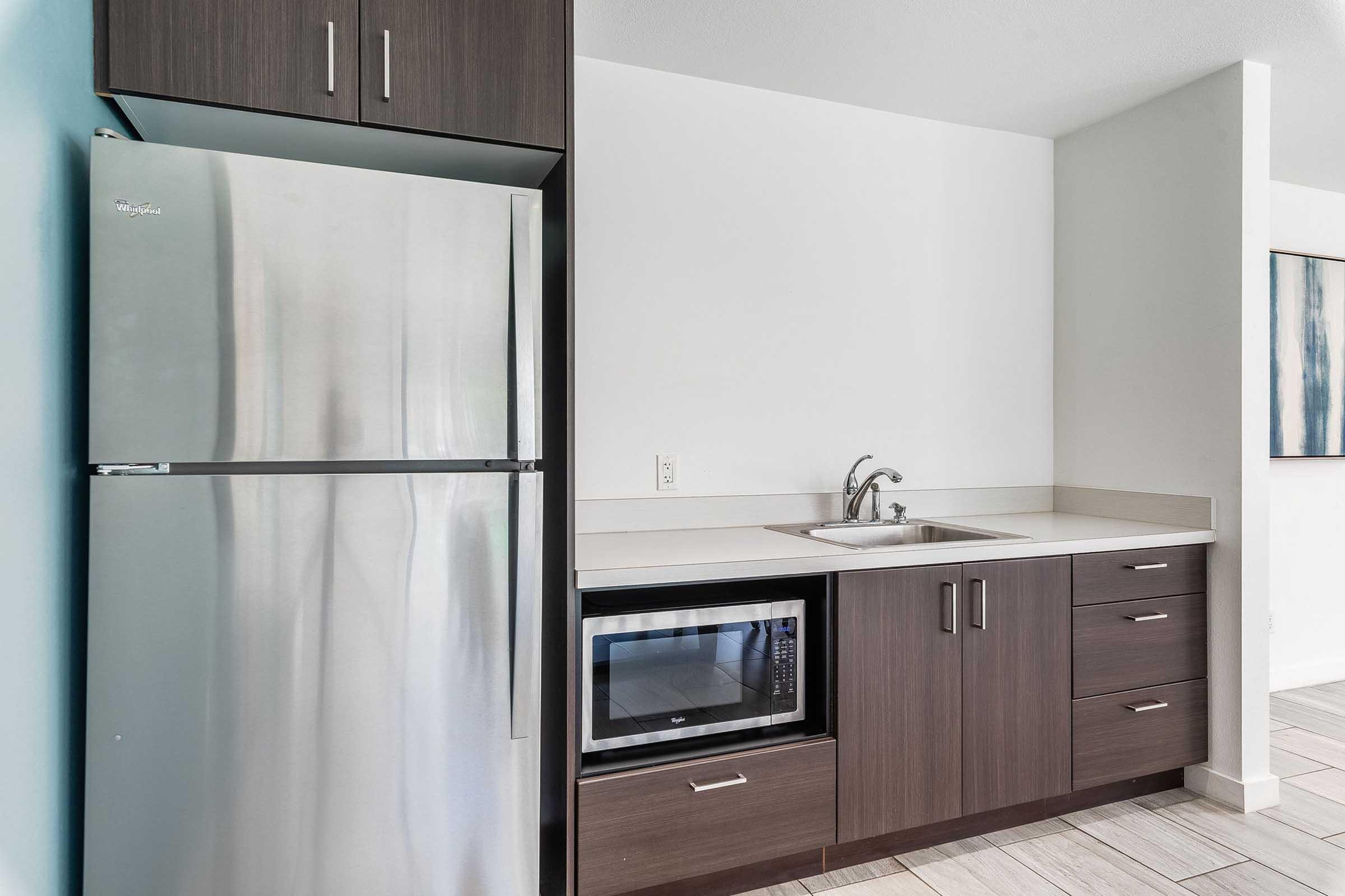 A modern kitchen featuring a stainless steel refrigerator, a microwave built into the cabinetry, and a sink with a sleek faucet. The cabinets are dark wood, providing contrast against the light-colored walls and countertops. The space is bright and clean, with tiled flooring.