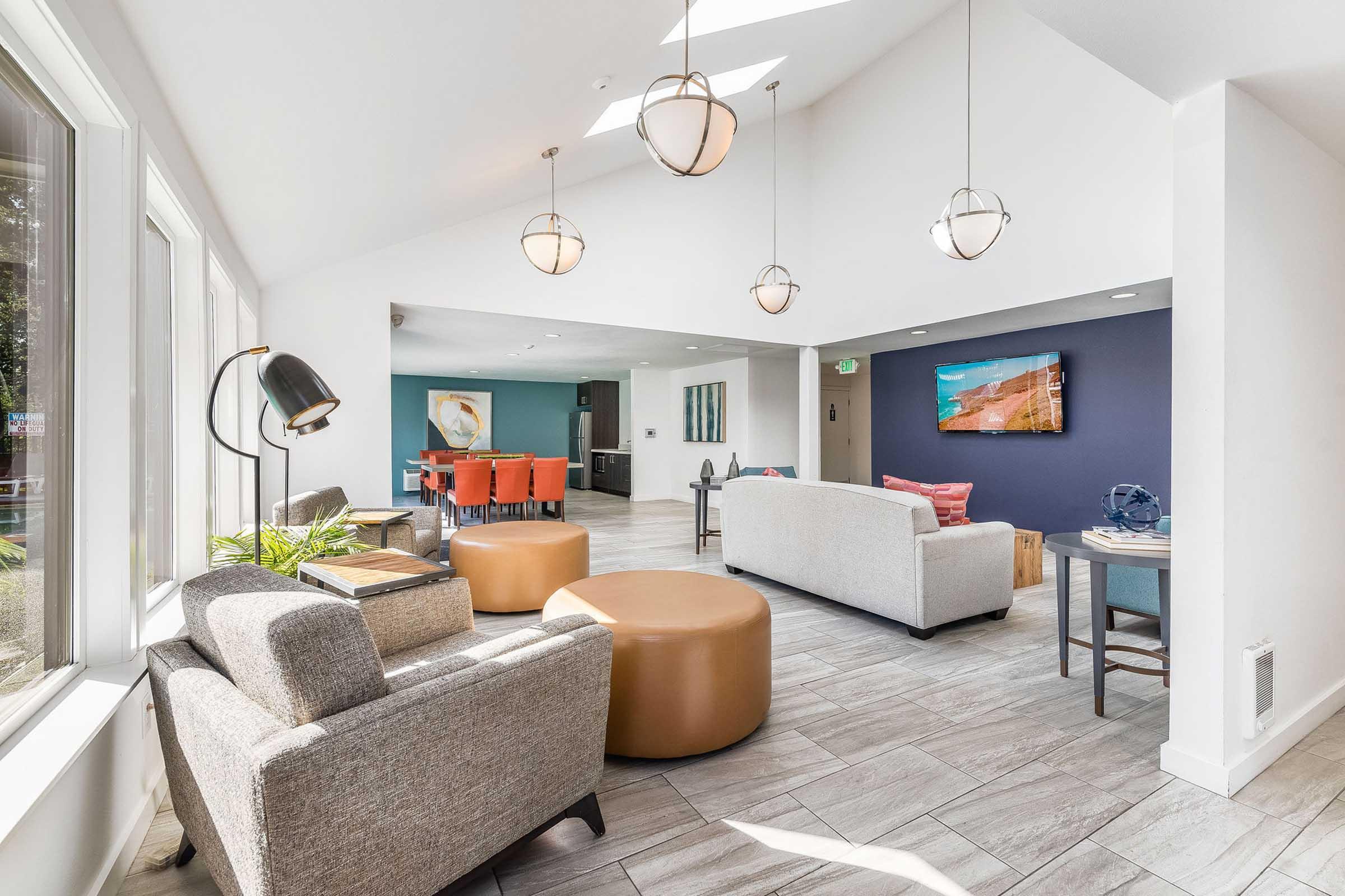 A bright, modern interior featuring a spacious lounge area with comfortable seating, including a gray sofa, a round coffee table, and stylish light fixtures. The space is decorated with colorful accent walls and large windows allowing natural light, creating a welcoming atmosphere.