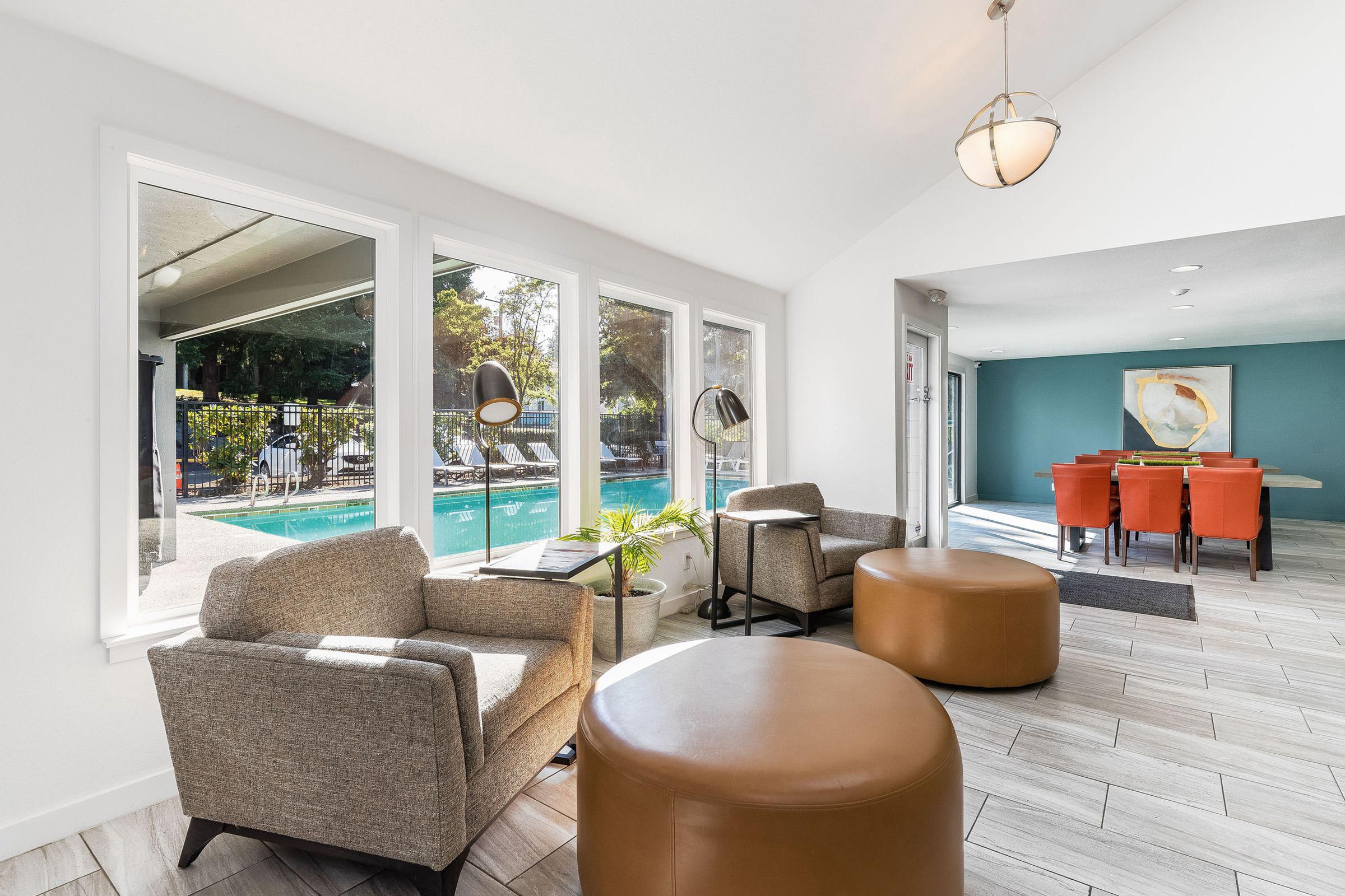 A cozy indoor lounge area featuring two gray armchairs and round ottomans, with large windows overlooking a pool area. A dining space with an orange table and chairs is visible in the background, along with potted plants and modern lighting fixtures. The decor combines comfort and modern style.