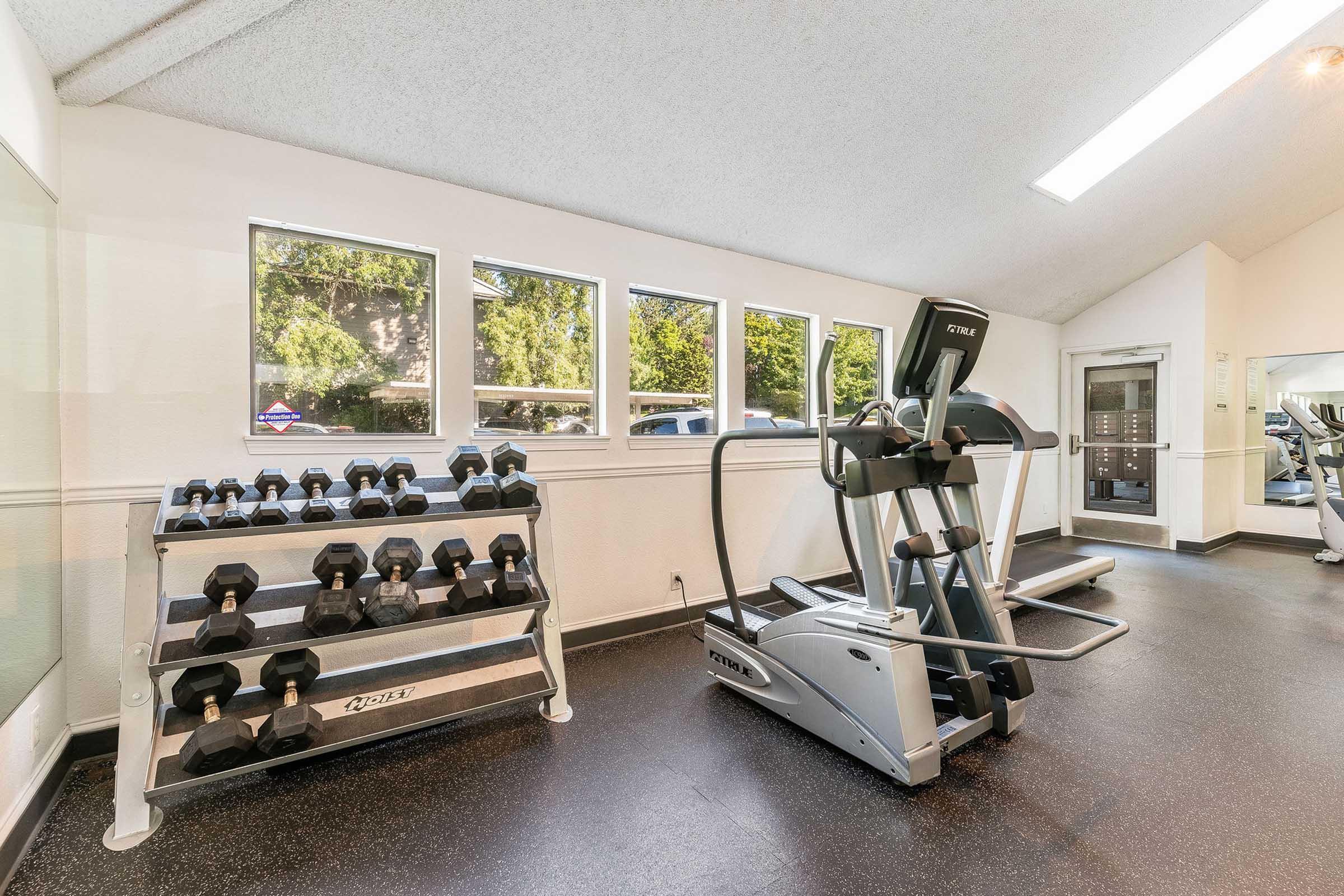 A modern gym interior featuring a rack of dumbbells, an elliptical machine, and large windows allowing natural light. The floor is covered in dark rubber flooring, and exercise equipment is well-maintained, creating a welcoming workout space.