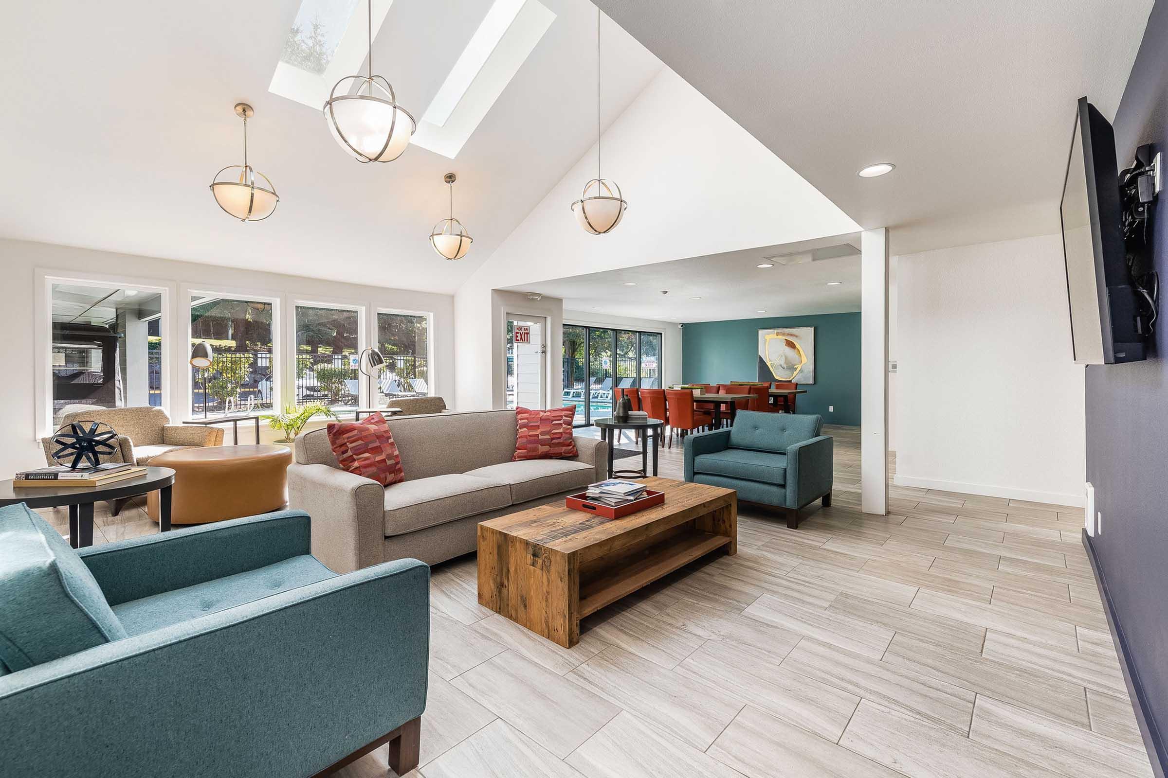 A modern lounge area featuring comfortable seating with two gray sofas, a turquoise chair, and a wooden coffee table. Large windows provide natural light, and pendant lights hang from the ceiling. A dining area with red chairs is visible in the background, along with a television mounted on the wall.