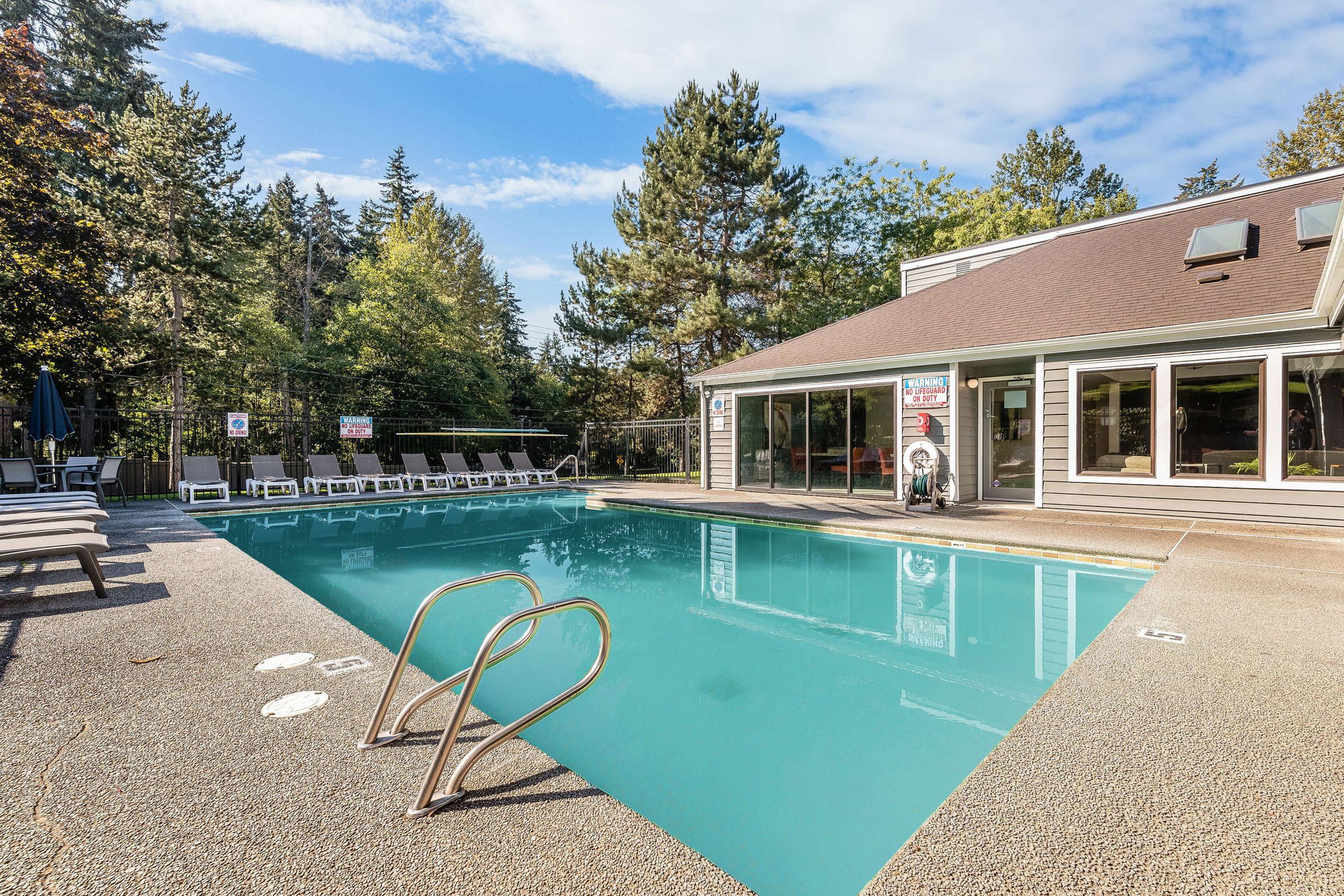 A clear blue swimming pool surrounded by lounge chairs, with trees in the background. A building with large windows is nearby, indicating a recreational area. The sky is partly cloudy, creating a serene atmosphere for relaxation and leisure activities.
