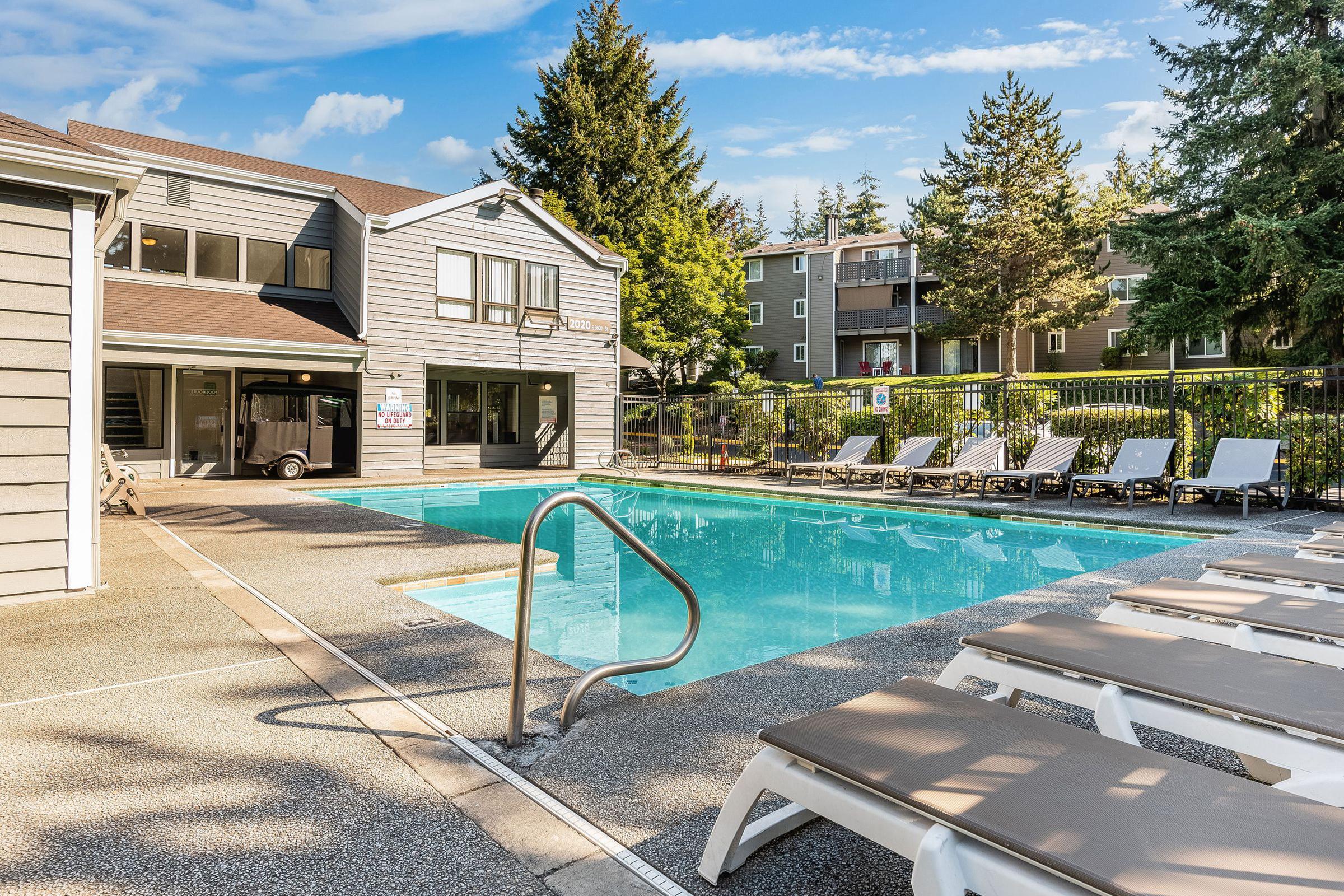 A sunny outdoor pool area featuring a clear blue swimming pool, surrounded by lounge chairs. In the background, there are apartment buildings and lush greenery. The setting conveys a relaxed and inviting atmosphere for leisure and recreation.