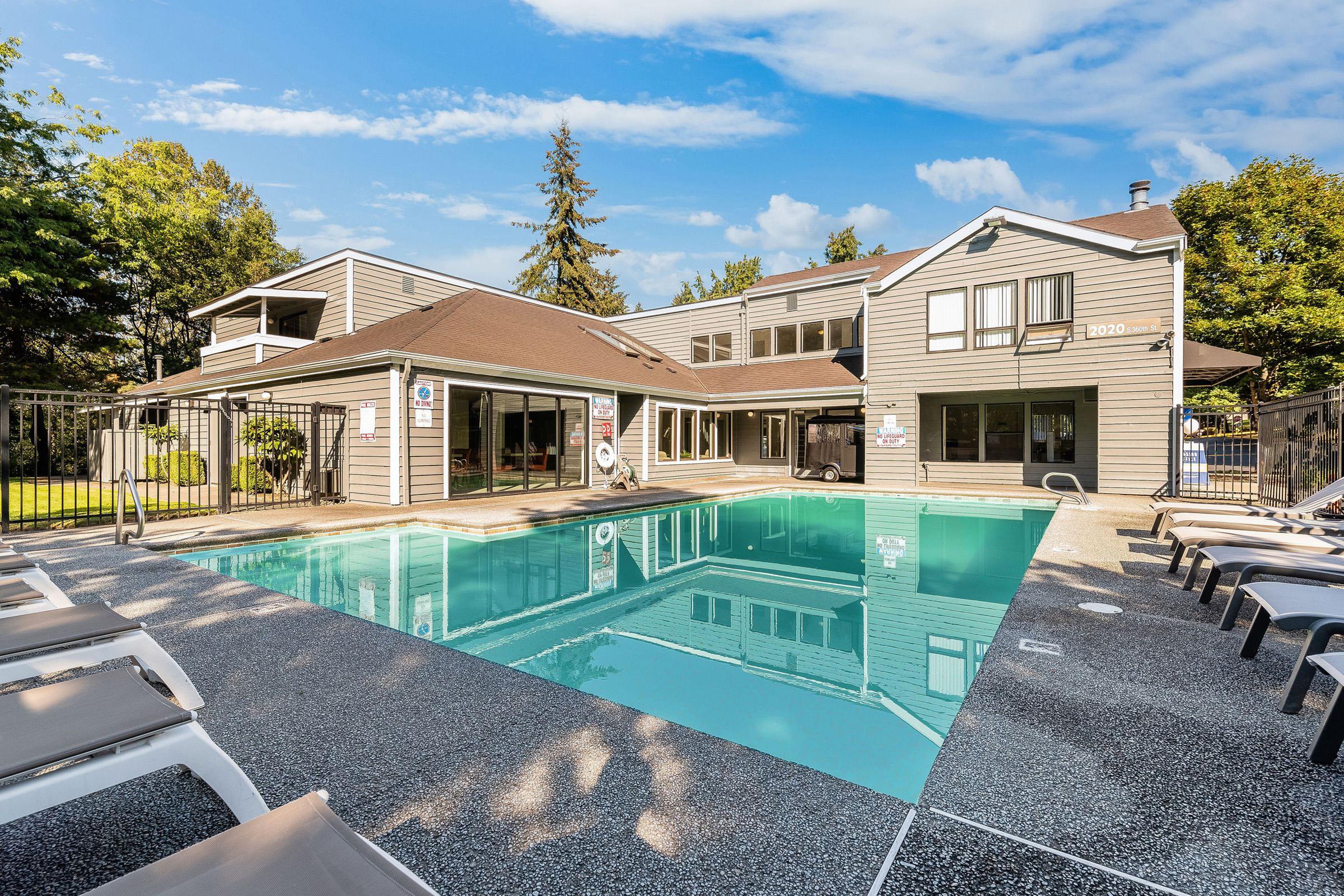 A clear blue pool surrounded by lounge chairs and a fenced area. The pool area is part of a modern building with large windows, green trees, and a sunny sky in the background. The scene conveys a relaxing outdoor atmosphere ideal for leisure activities.