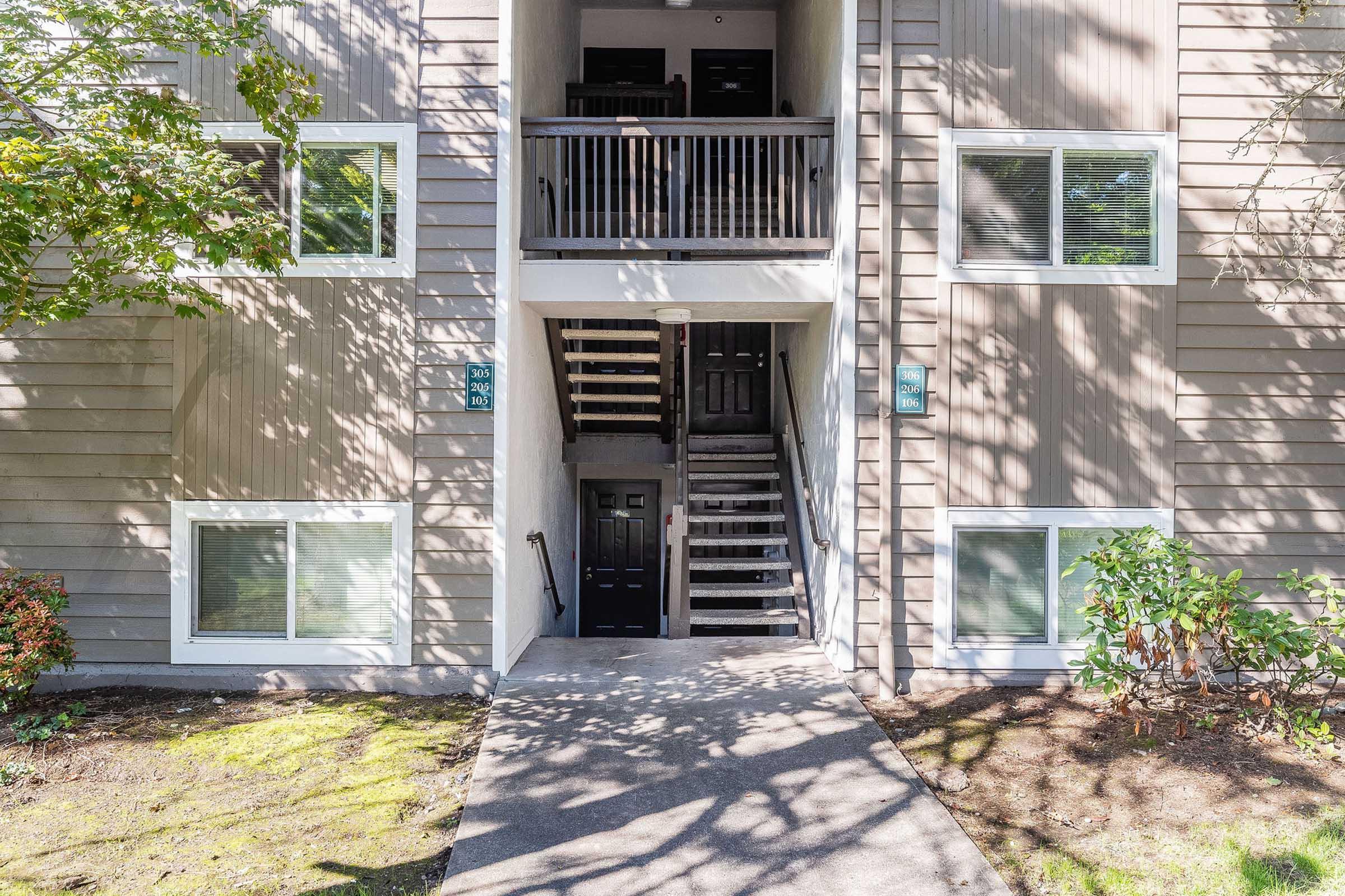 Exterior view of an apartment building featuring a set of black double doors at the entrance and a staircase leading to the upper level. The building has light gray siding, with several windows and landscaped greenery around the entrance pathway. Sunlight casts shadows on the facade.