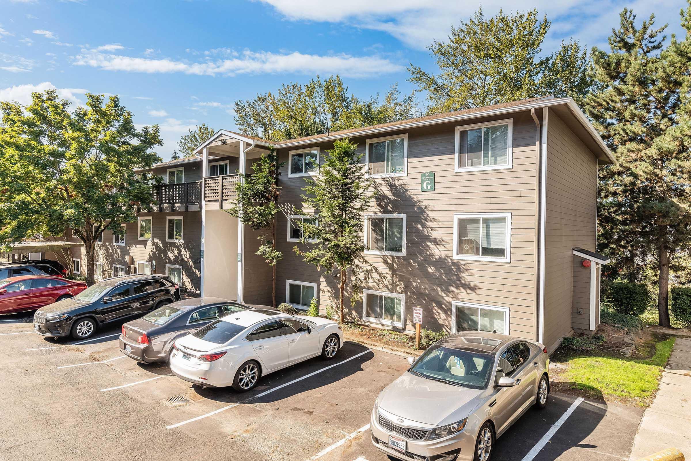 A multi-story apartment building surrounded by trees, with a parking lot in front containing several cars. The building features multiple windows and a balcony on the upper floor. The sky is clear with a few clouds, indicating a sunny day.