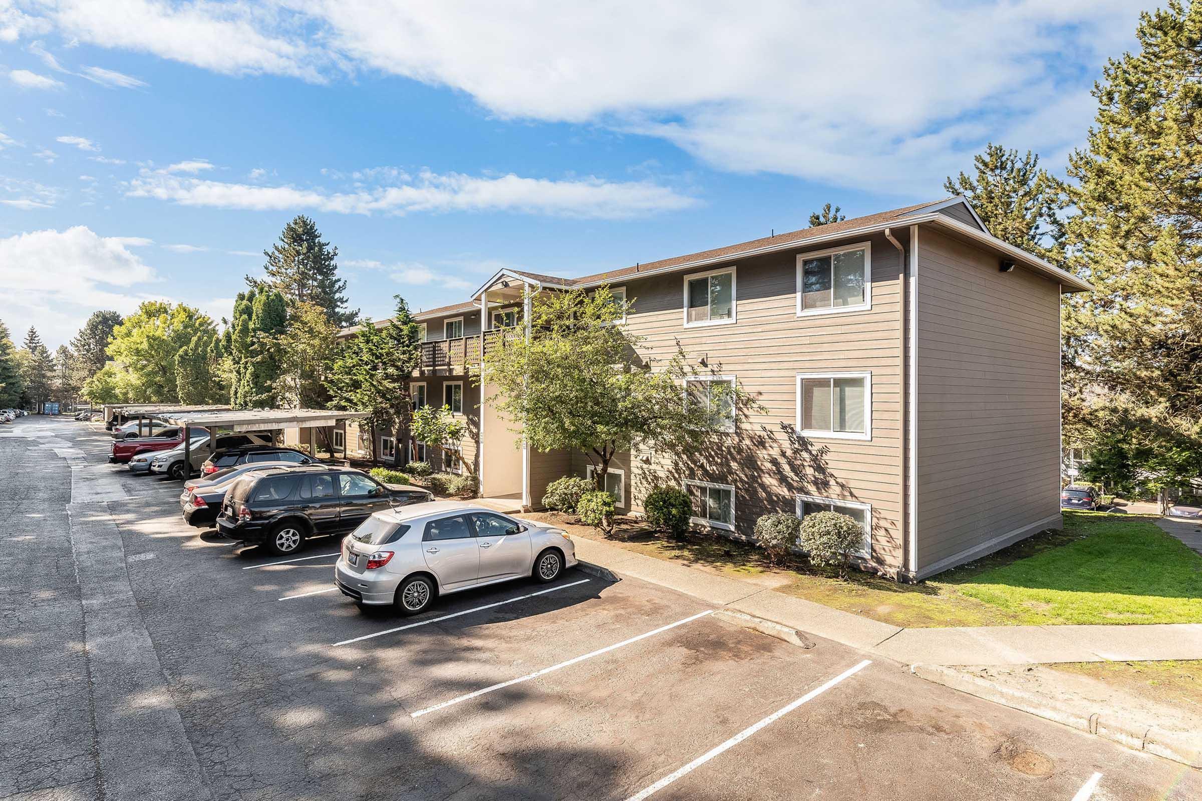 An apartment building with two stories, surrounded by trees and grass. Several parked cars are visible in the foreground, and the sky is partly cloudy. The building features wooden siding and windows, with a paved parking area and a well-maintained landscape.