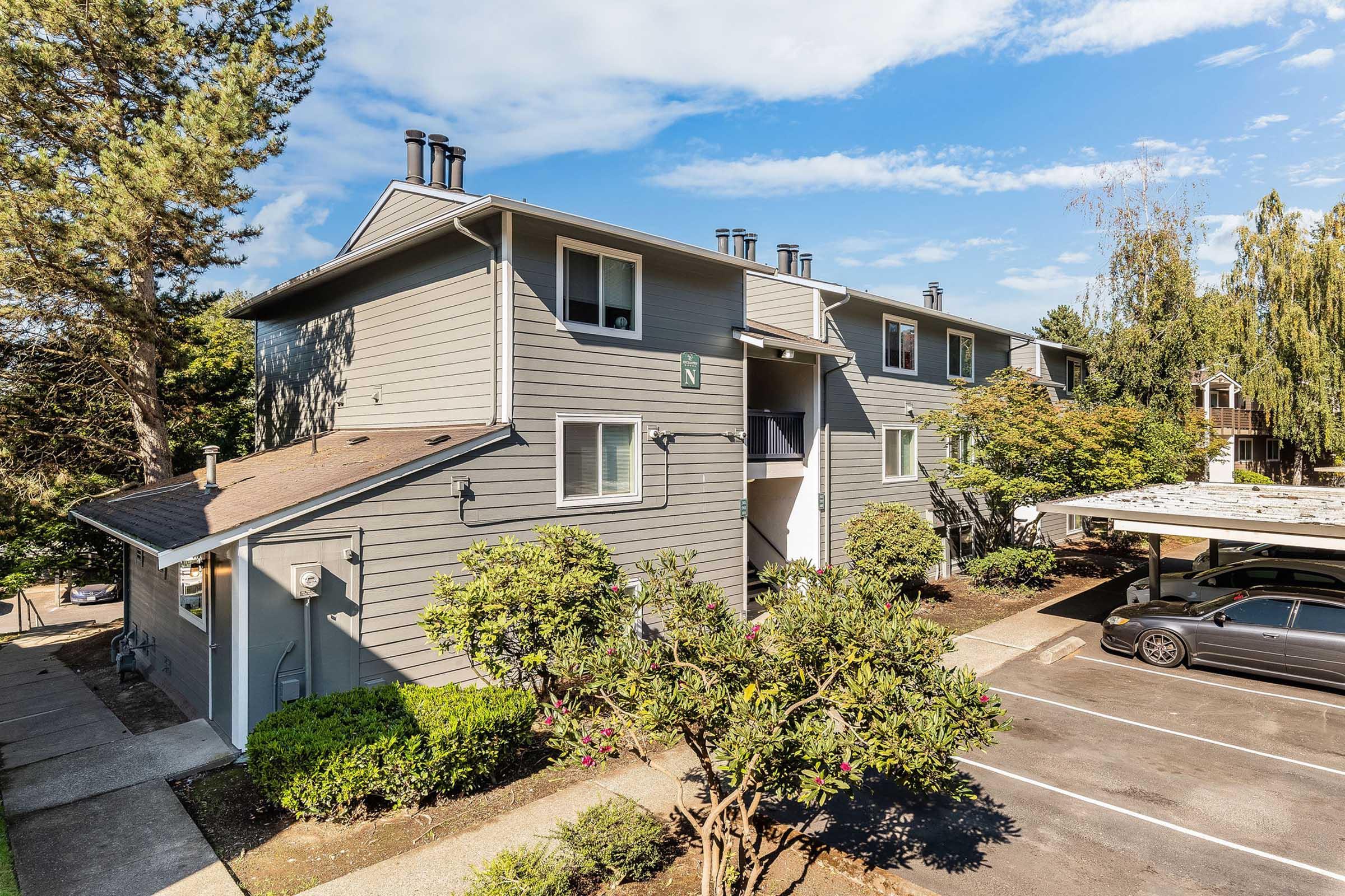 Exterior view of a multi-unit residential building with gray siding. The building has multiple balconies and windows, surrounded by landscaping with trees and shrubs. A parking area is visible in the foreground, with parked cars under a shaded structure. Bright blue sky with scattered clouds in the background.