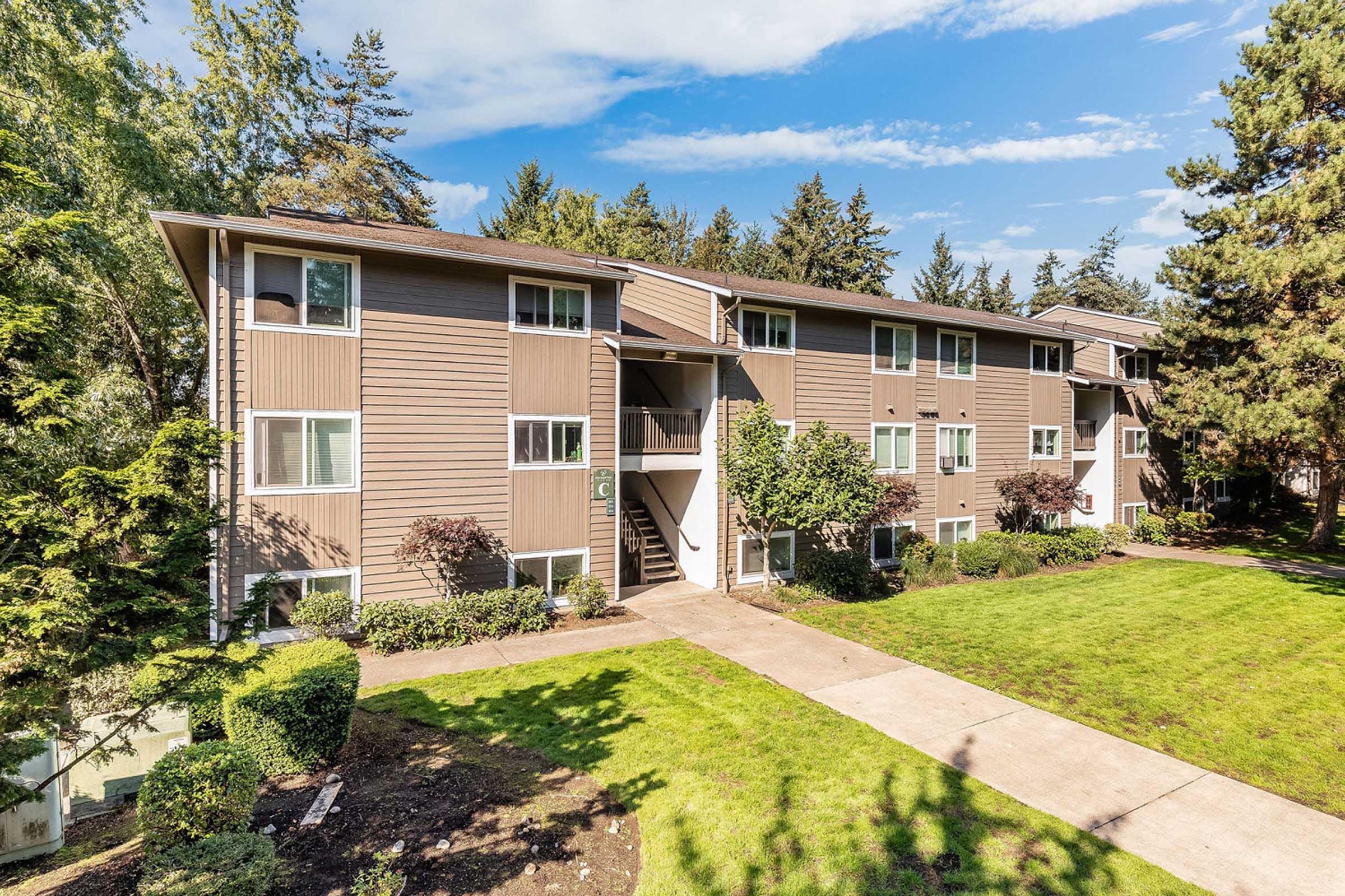 A three-story apartment building with a light brown exterior, surrounded by green grass and trees. The entrance features a staircase leading to the upper floors, and there are neatly trimmed bushes along the pathway. The sky is clear with a few clouds, creating a pleasant atmosphere.