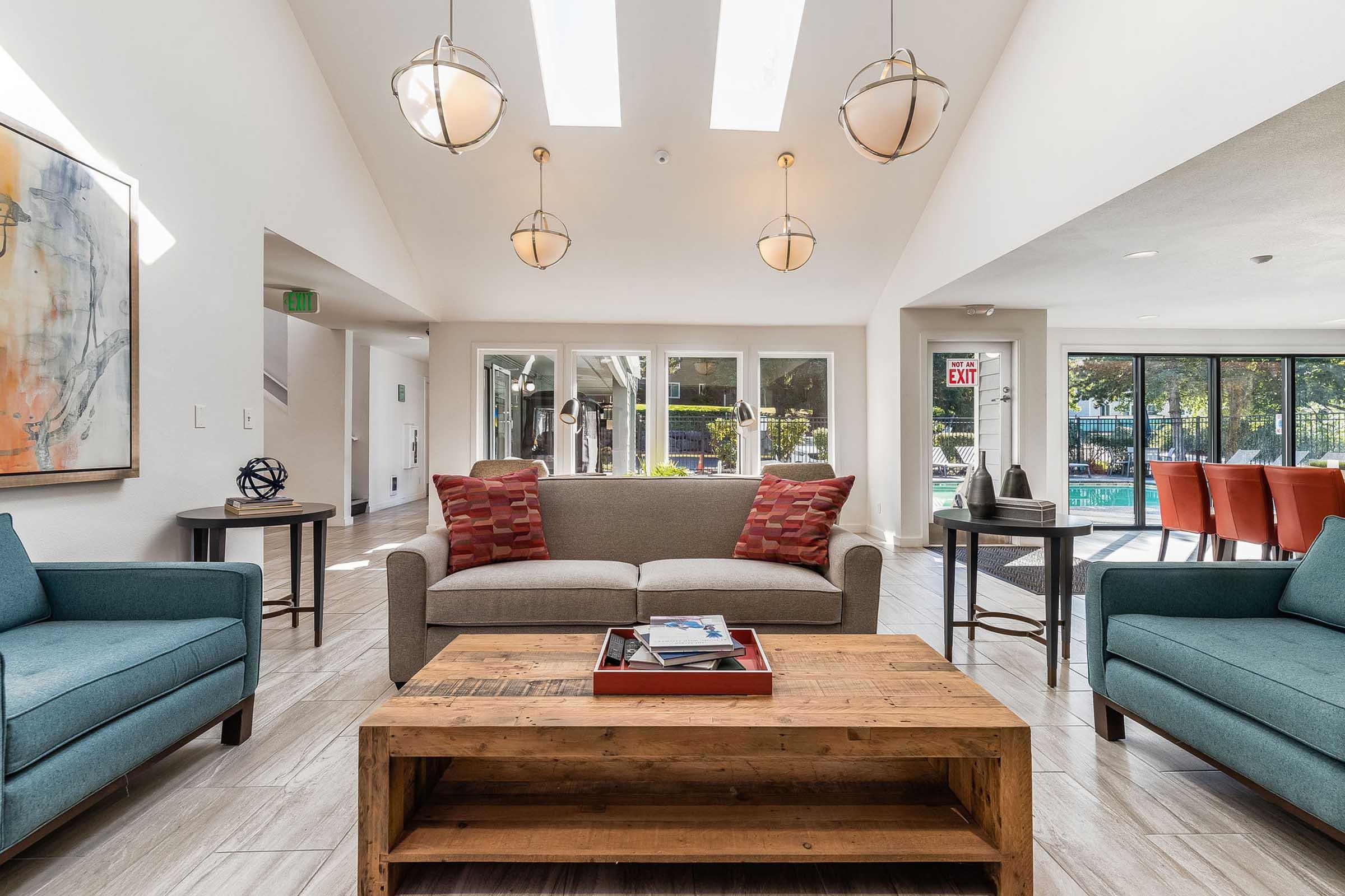 A modern lounge area featuring a gray sofa adorned with red accent pillows, two teal armchairs, and a rustic wooden coffee table displaying magazines. The space is illuminated by pendant lights and has large windows with a view of an outdoor area. The walls are painted white, creating a bright and welcoming atmosphere.