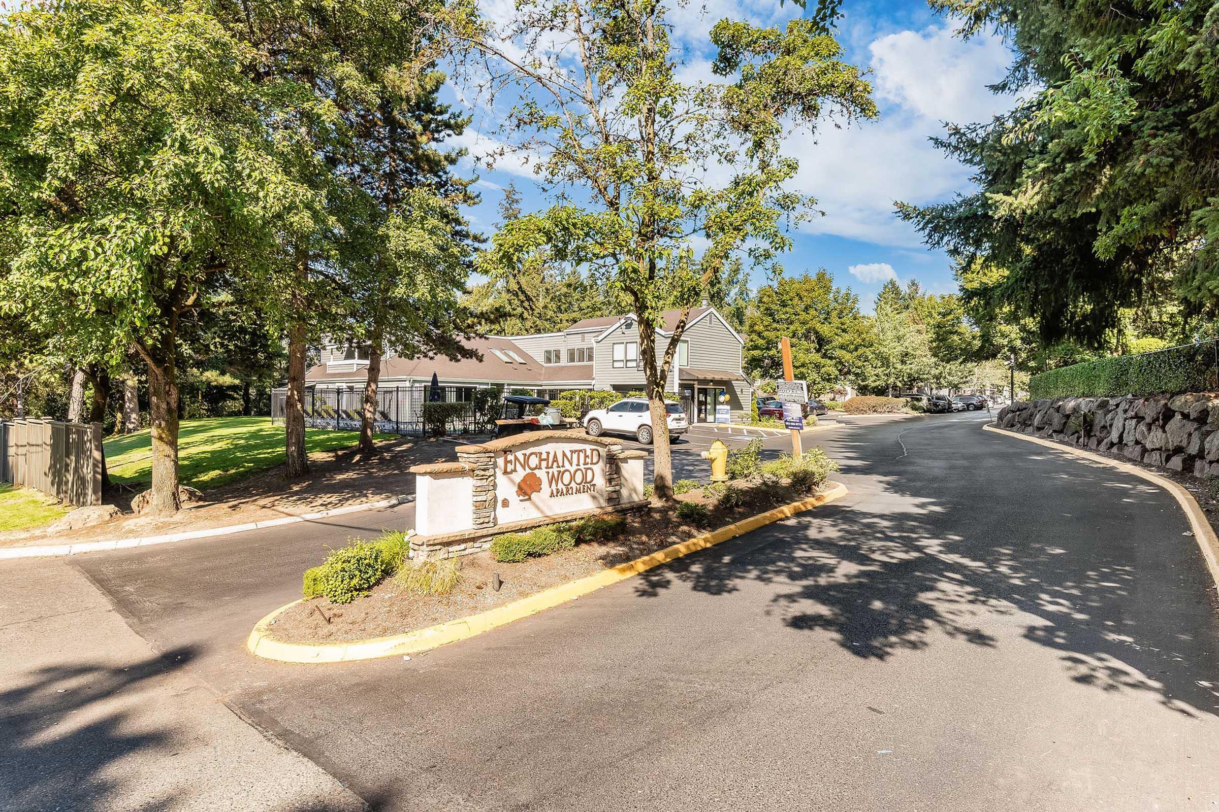 Entrance to Enchanted Woods, featuring a sign with the name prominently displayed. The area is surrounded by lush greenery, trees, and a clear blue sky. A paved road leads into the property, with parked vehicles visible in the background.