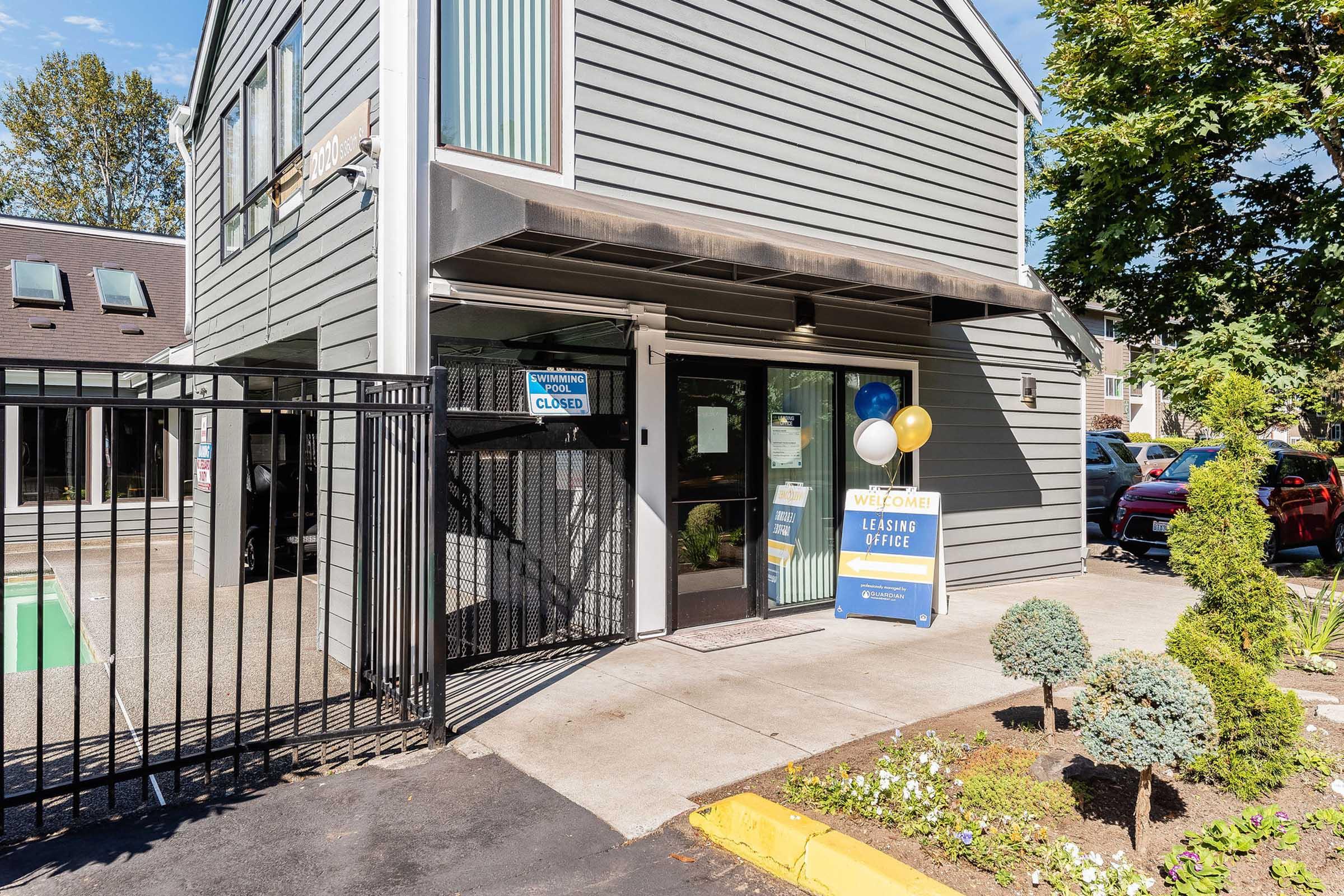 A leasing office entrance with a gated access, featuring a sign that reads "Leasing Office Closed." The building is gray with large windows, and there are decorative plants and balloons near the entrance. A pool area is visible in the background. Bright daylight enhances the scene.