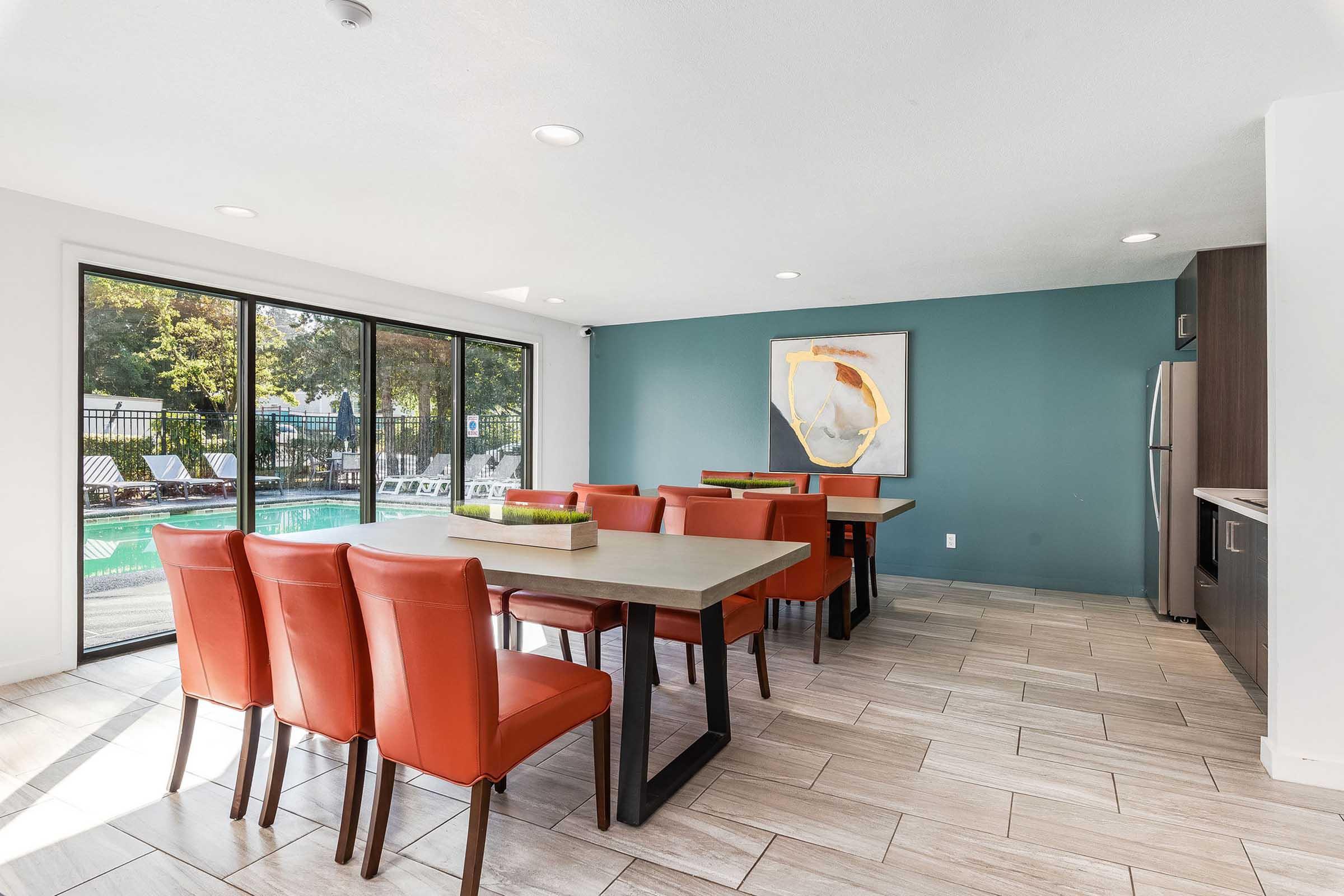 A modern dining area featuring several orange chairs around large rectangular tables, with large windows overlooking a swimming pool. The walls are painted a soothing teal, and there is a piece of abstract art on one wall. Bright lighting enhances the airy atmosphere of the space.