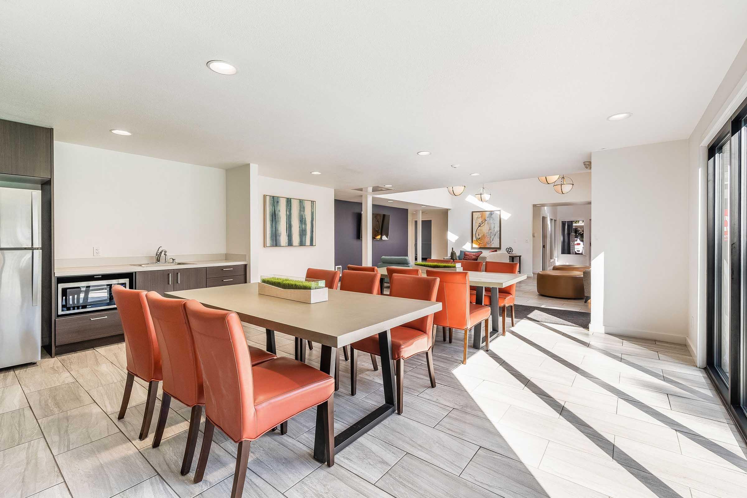 A modern dining area with a large table surrounded by orange chairs. There is a kitchenette featuring a refrigerator and sink. Natural light fills the space through large windows, highlighting the contemporary decor and neutral color palette. The room is open and airy, creating a welcoming atmosphere.