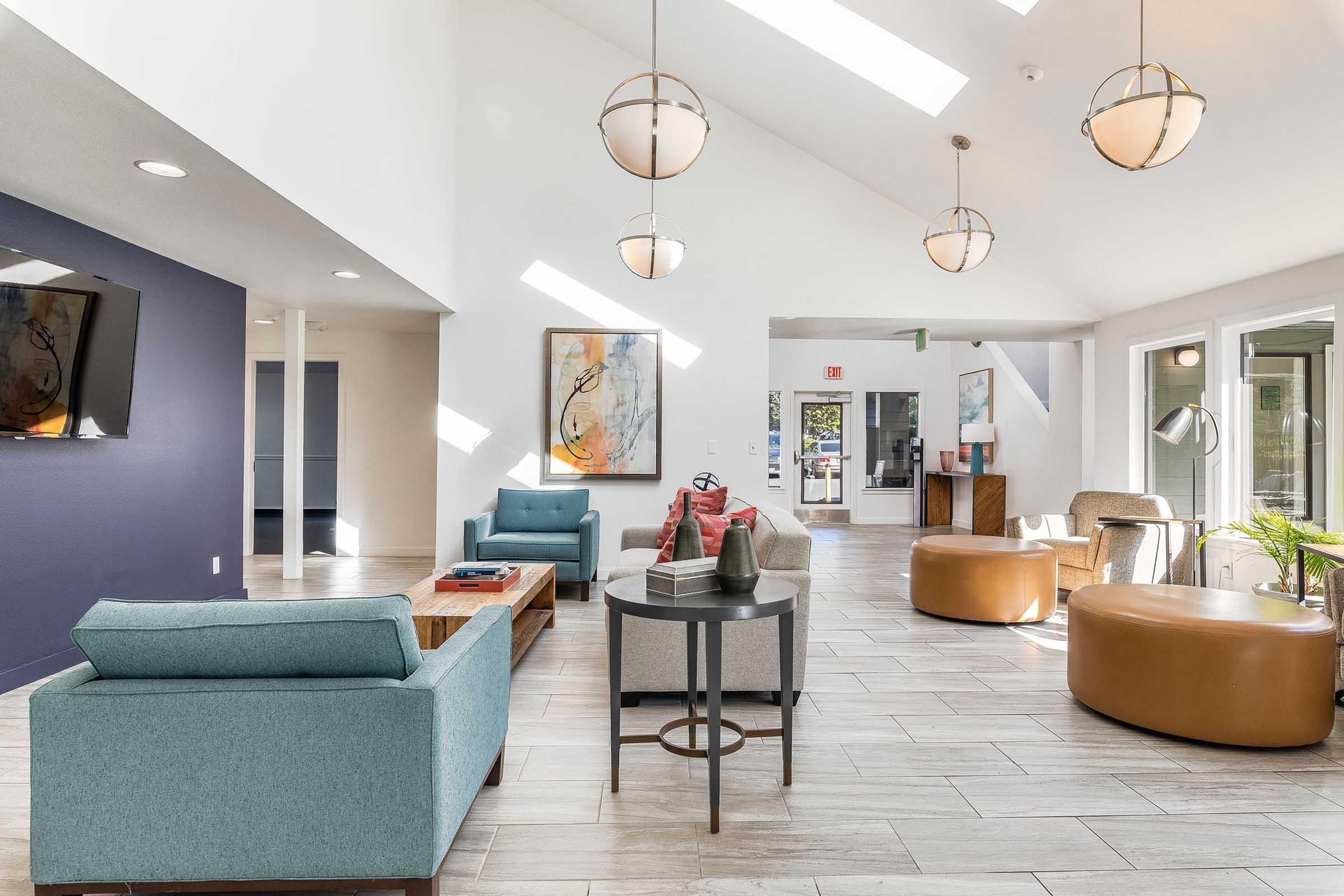 A modern, well-lit lobby featuring a mix of stylish seating, including a blue sofa, gray chairs, and brown poufs. The walls are painted white and dark blue, adorned with abstract art. Large windows allow natural light to flood the space, creating a welcoming atmosphere.
