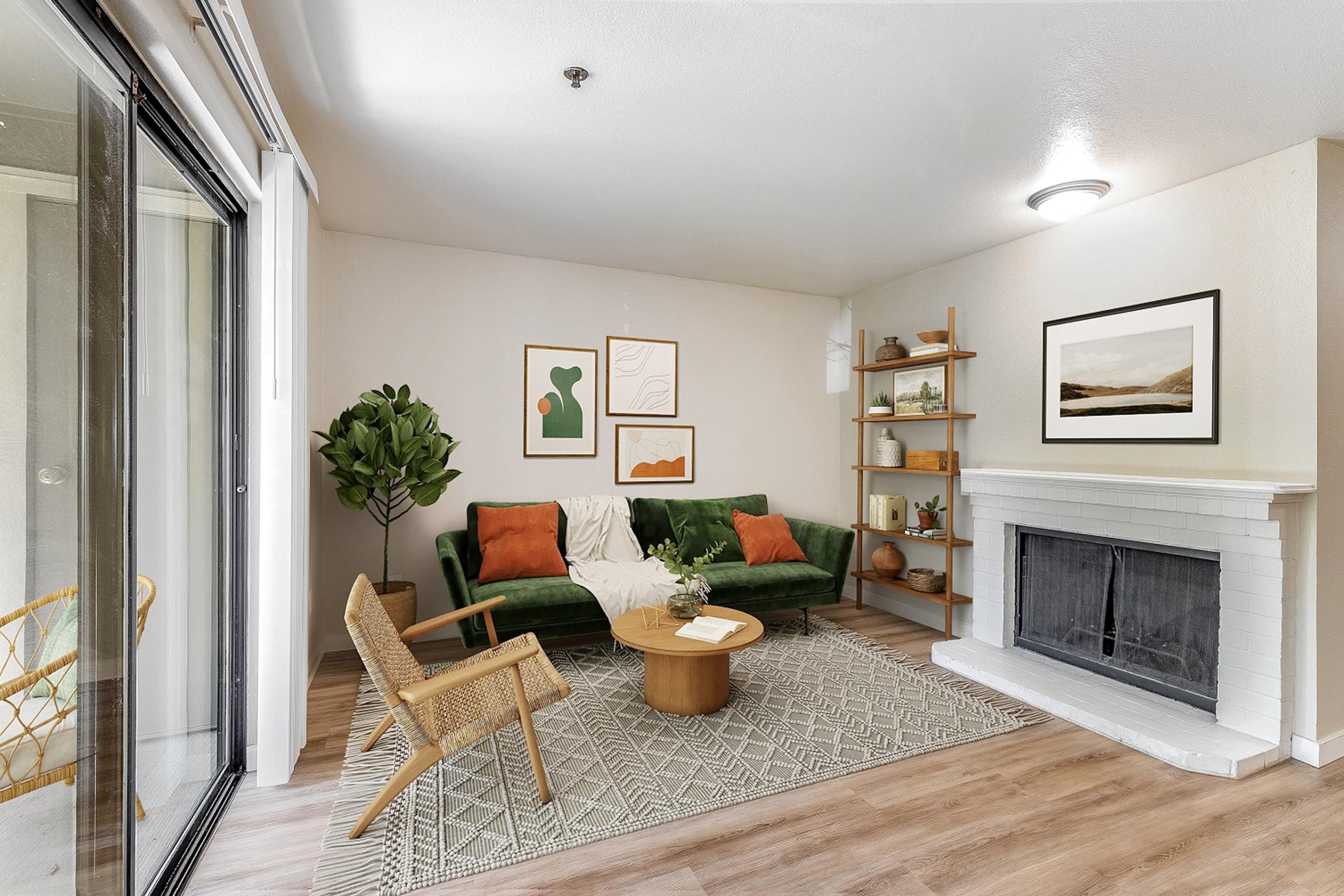 A cozy living room featuring a green sofa with orange and green cushions, a round wooden coffee table, and a woven rug. There are framed artworks on the wall, a shelf with decor items, a potted plant, and a fireplace. Natural light streams in through large sliding glass doors.