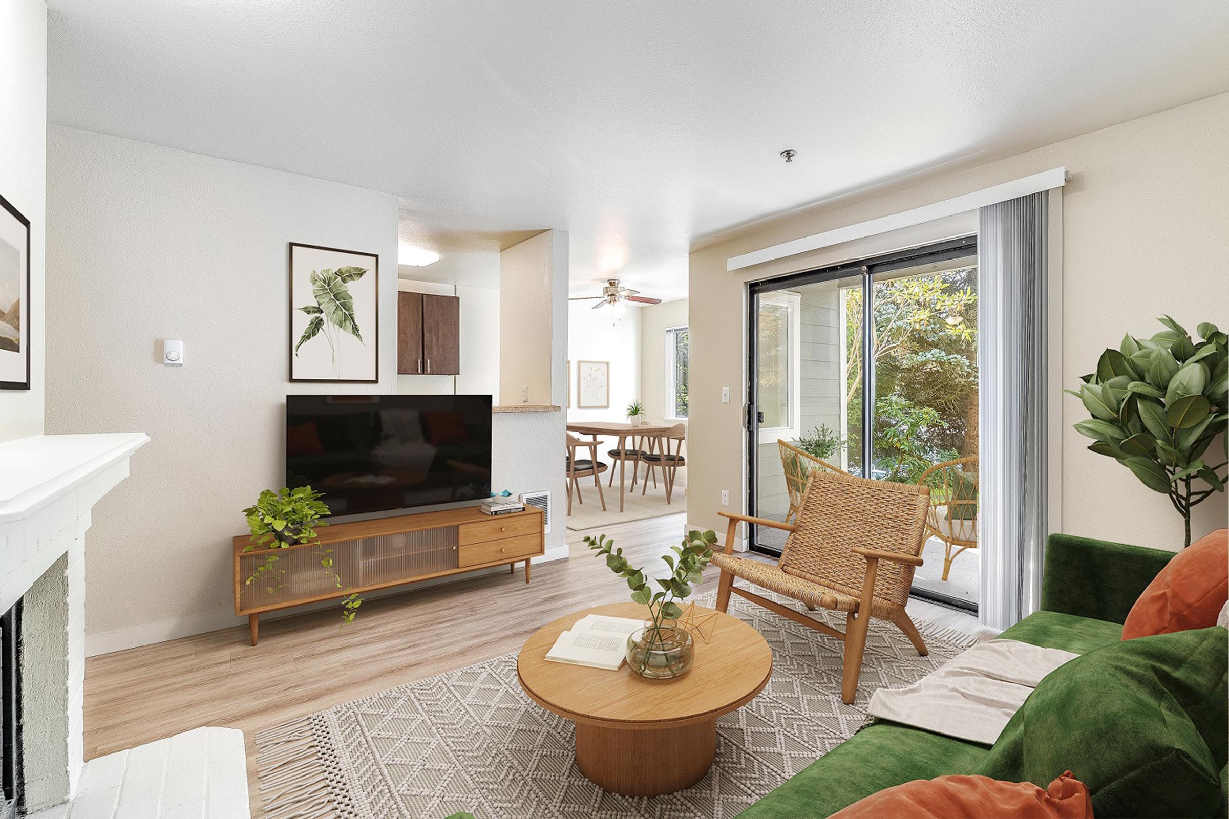 A cozy living room with a green sofa, a wooden coffee table, and a TV on a mid-century modern console. Large windows allow natural light, revealing a view of a small garden. There are framed art pieces on the walls, a ceiling fan, and a dining area visible in the background, creating a warm and inviting atmosphere.