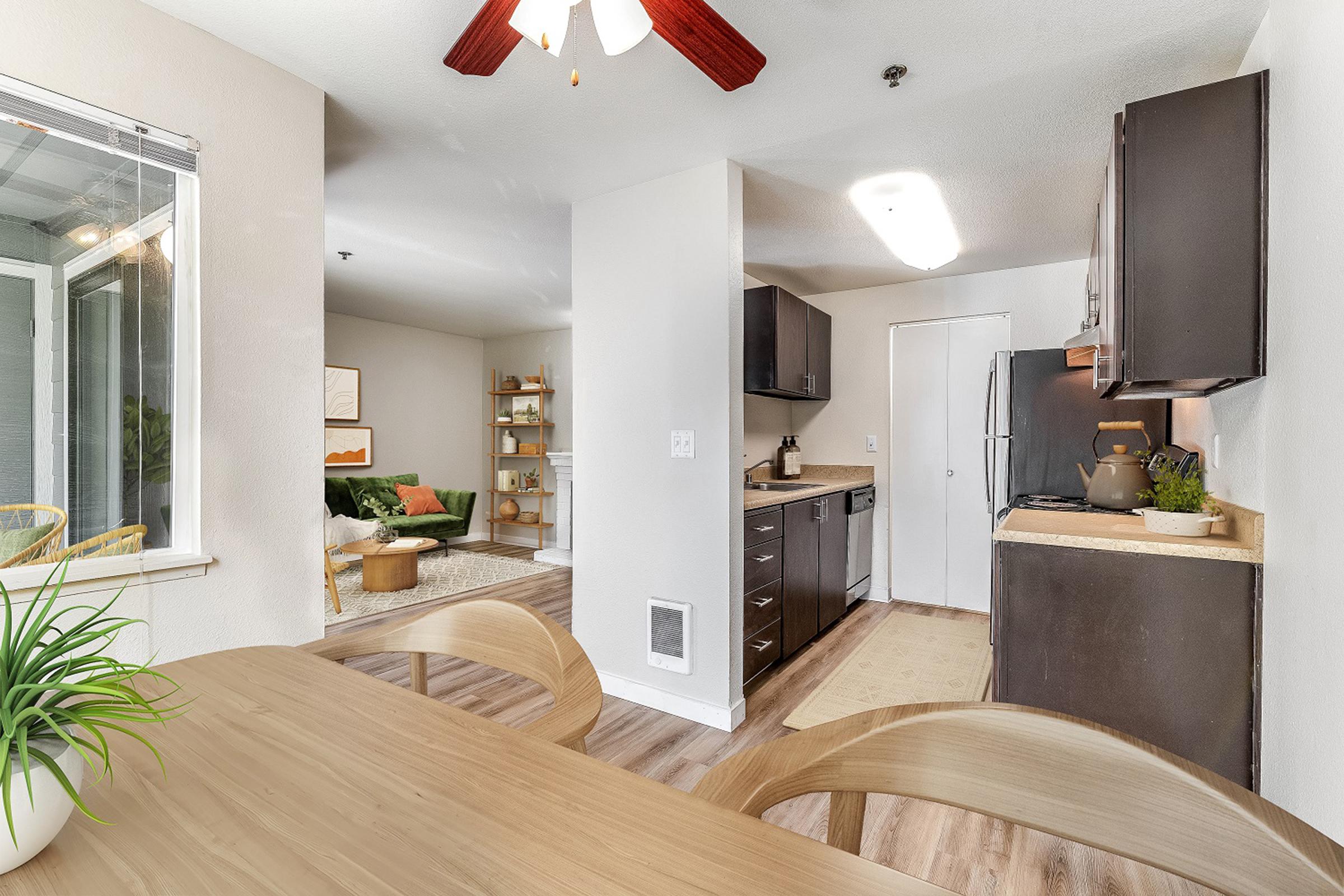 A modern kitchen and dining area with wooden table and chairs. The kitchen features dark cabinets, a stove, and a refrigerator. Through an archway, a cozy living space with a green sofa and bookshelves is visible, along with natural light coming from a window. The decor is light and inviting.