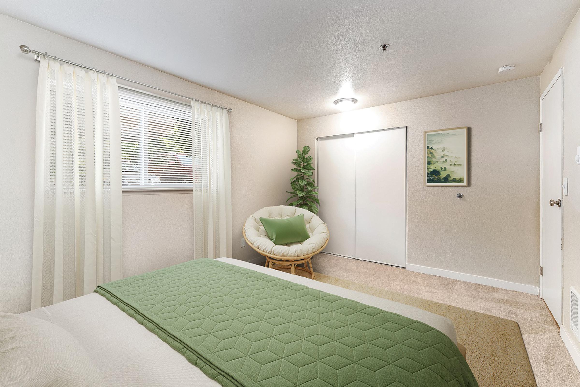 A cozy bedroom featuring a green bedspread, a round chair with a green cushion, and a small plant. Natural light enters through a window adorned with sheer curtains. A closet is visible with sliding doors, and a framed picture hangs on the wall, adding a touch of decor. The carpeted floor enhances the warm ambiance.