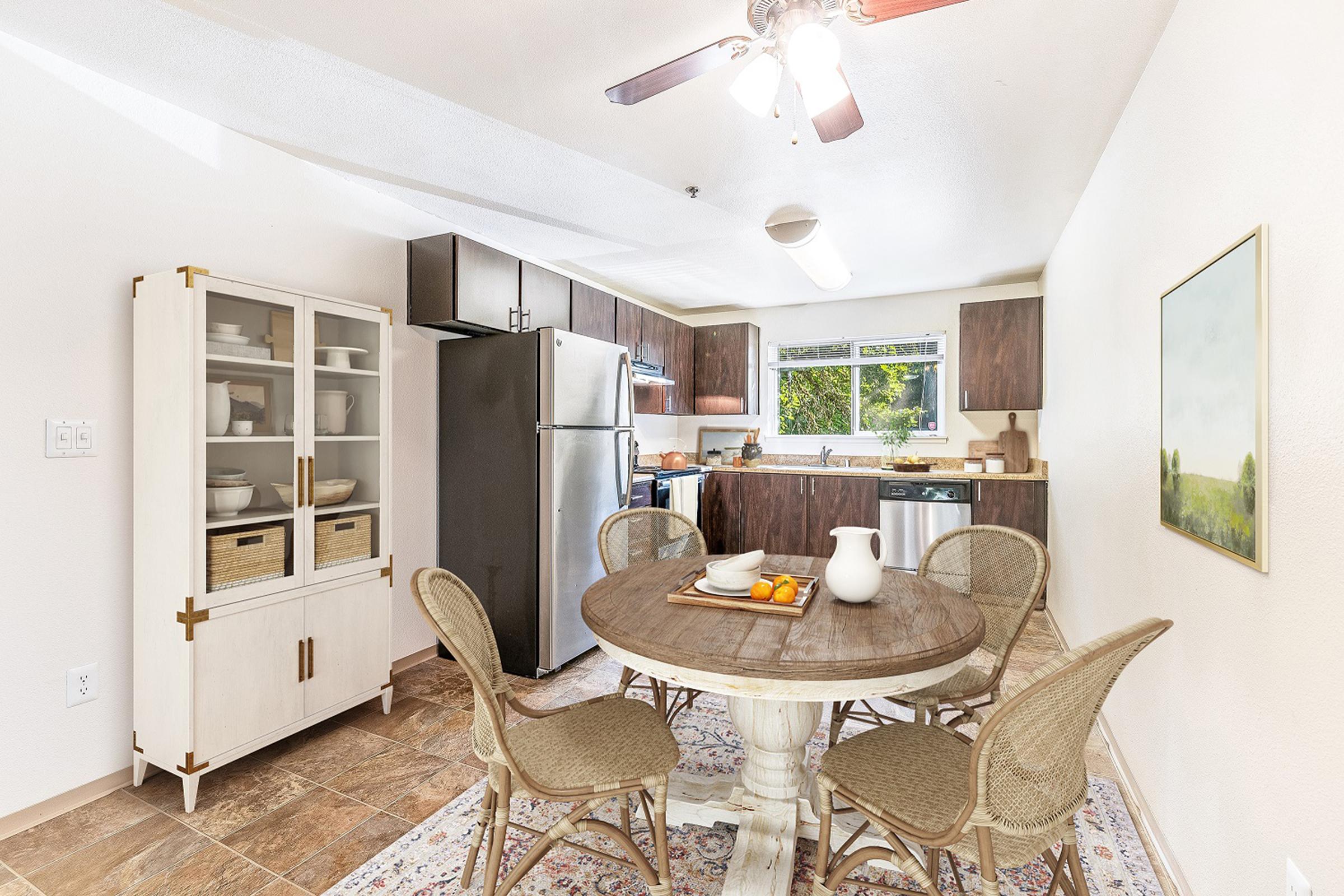 Bright kitchen featuring a round table with four chairs, a white cabinet, and stainless steel appliances. The cabinets have a dark wood finish, and there’s a window providing natural light. The table is set with a pitcher and fruit. The floor is tiled, and there's a decorative rug underneath the table.