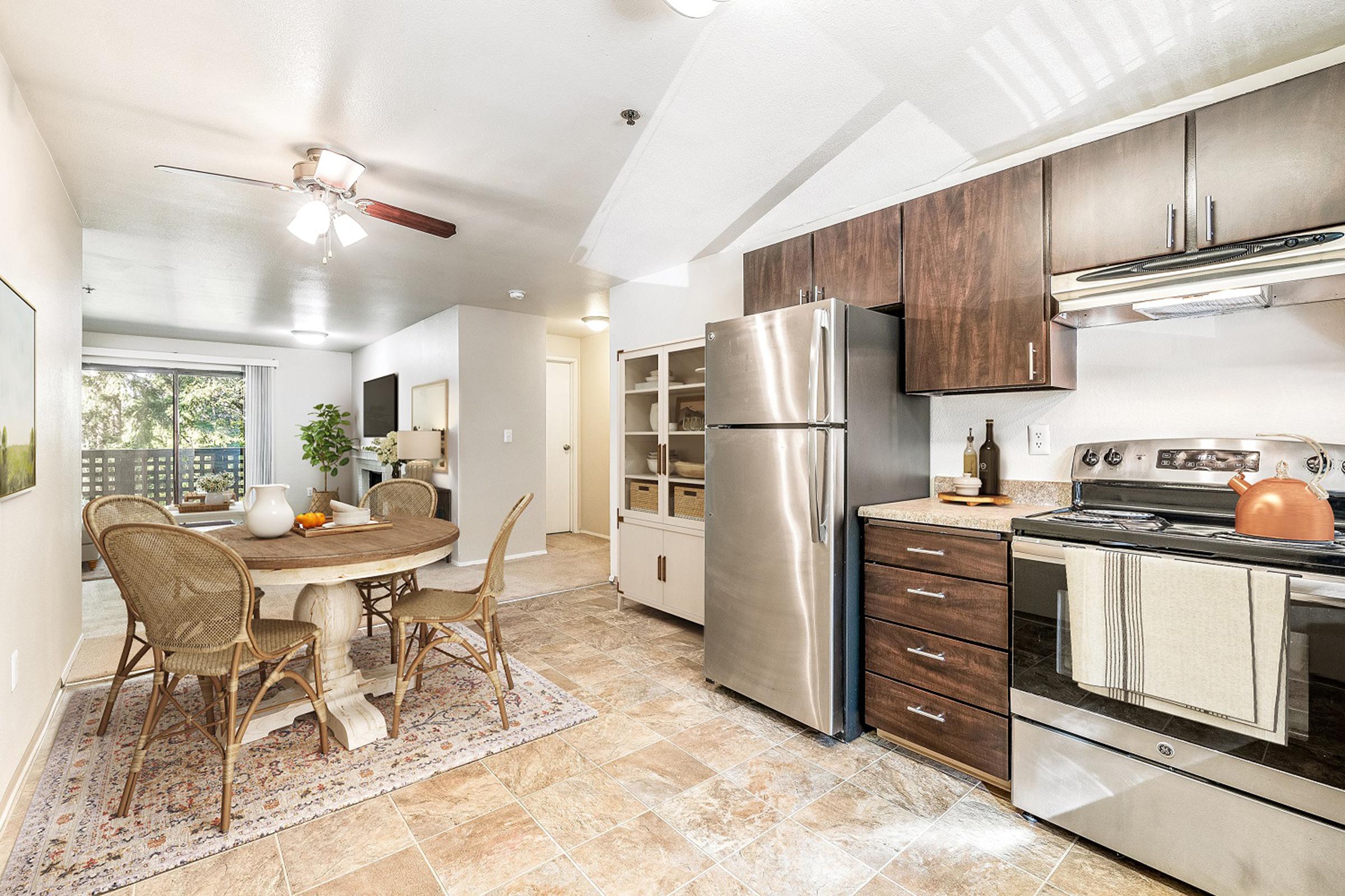 A modern kitchen and dining area featuring stainless steel appliances, wooden cabinets, a round table with wicker chairs, and a decorative rug. Natural light streams in from sliding glass doors that open to a patio, creating a warm and inviting atmosphere.