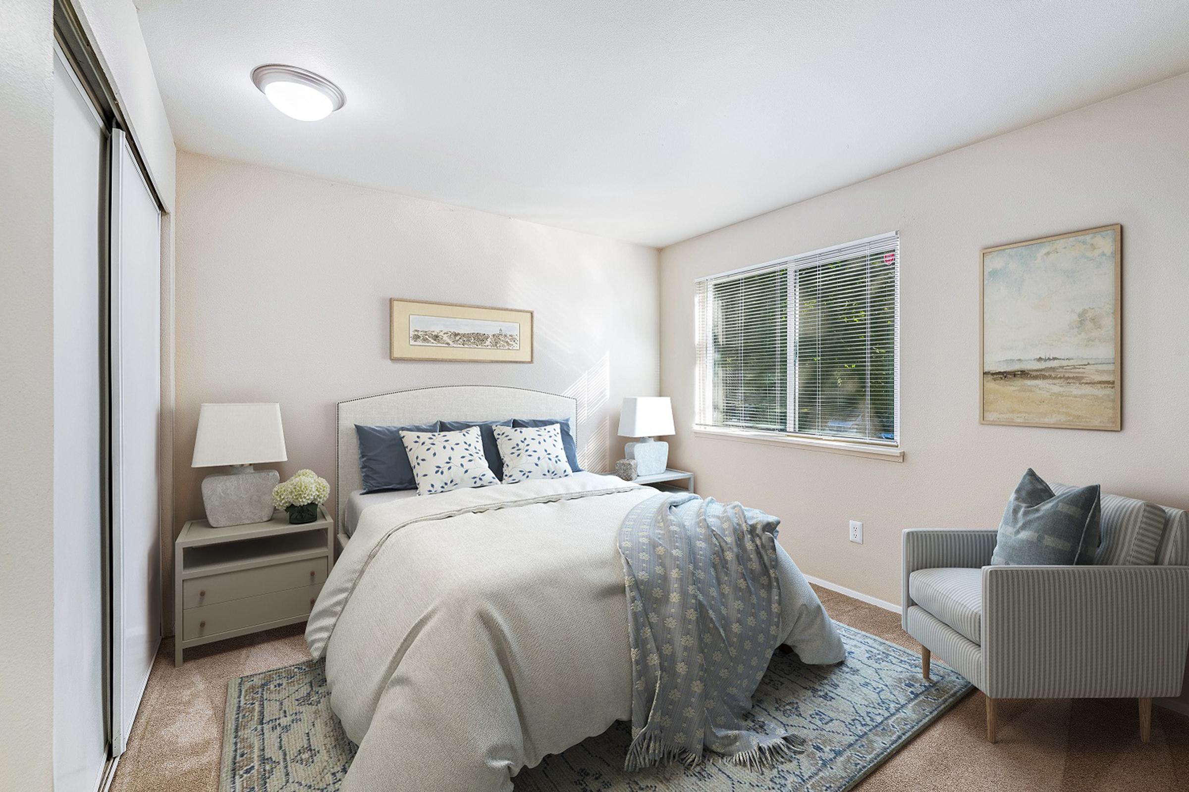 Cozy bedroom featuring a queen-sized bed with light blue and white bedding, two bedside lamps on nightstands, a comfortable armchair, and a window with natural light. The walls are painted in soft beige, and there is a decorative rug on the carpeted floor. Artwork hangs above the bed.