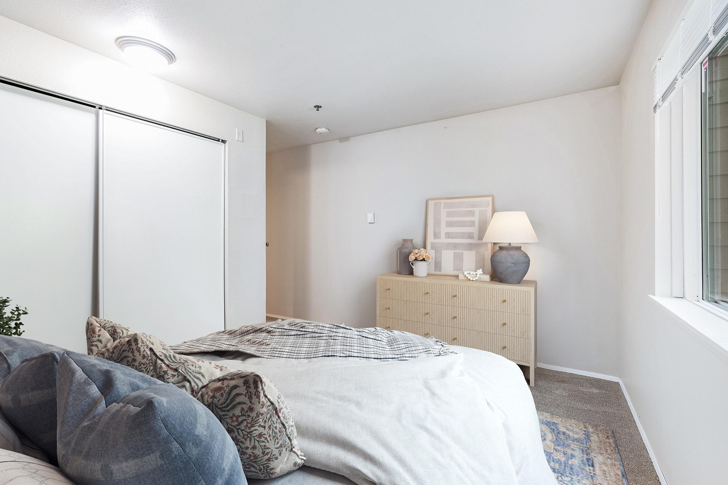 A cozy bedroom featuring a neatly made bed with decorative pillows, a light-colored dresser, a bedside lamp, and a window allowing natural light. The room has a neutral color palette, a soft rug, and minimal decor, creating a calm and inviting atmosphere.