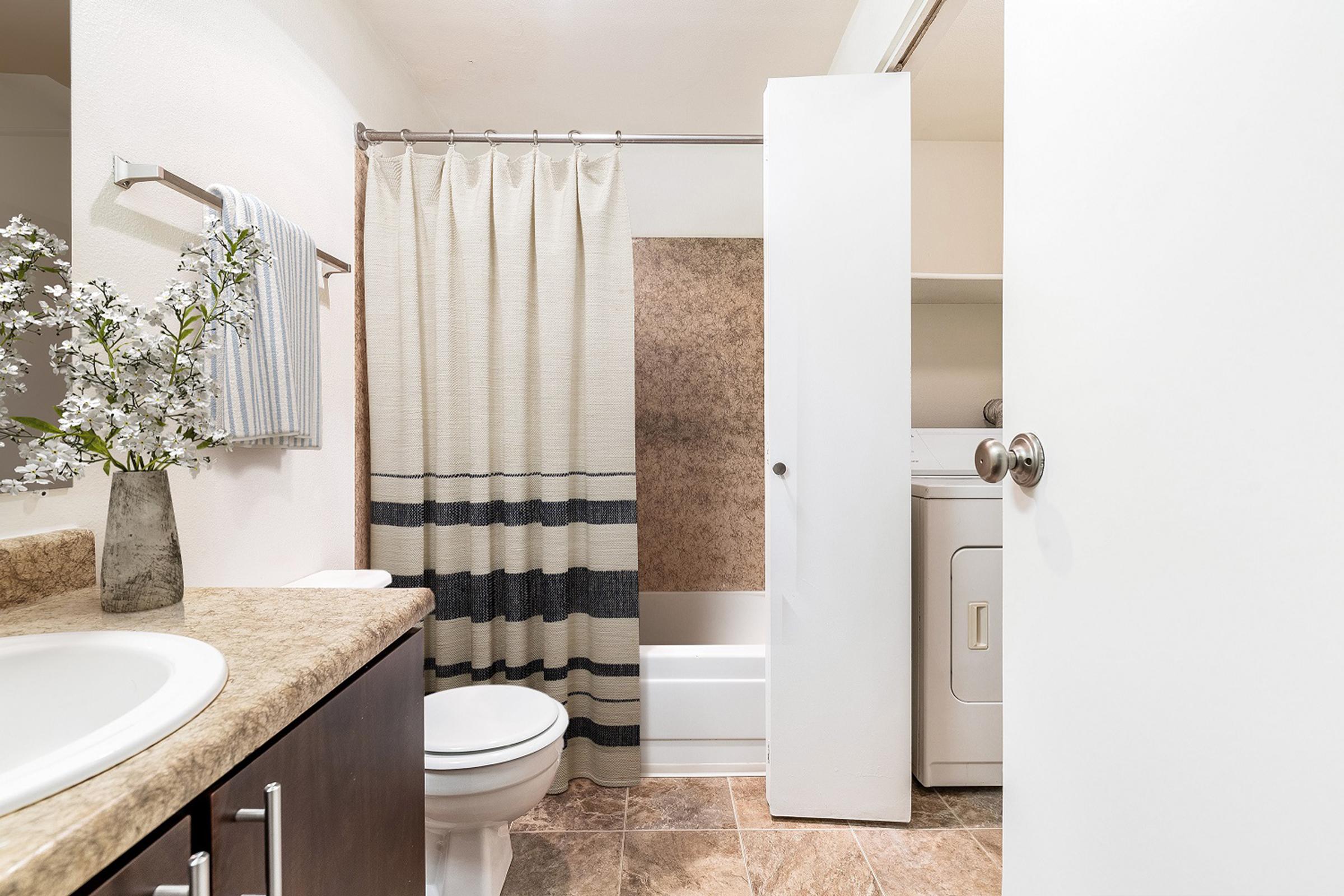 A clean bathroom featuring a white bathtub with a shower curtain, a toilet, and a sink with a beige countertop. A laundry area is visible near the back. The decor includes a vase with flowers and striped towels hanging on a rack. The walls are light-colored, creating a bright and open atmosphere.