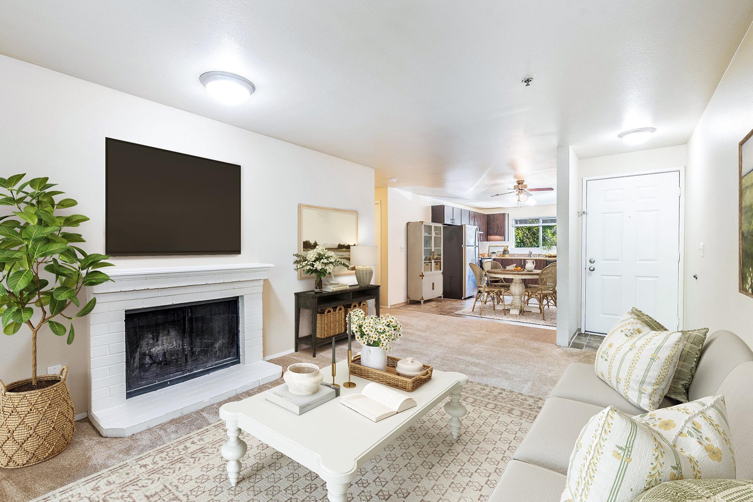 A modern living room featuring a light-colored sofa with decorative pillows, a coffee table with decorative items, and a fireplace. A large TV mounted on the wall is visible. In the background, there's a dining area with a table and chairs, and a door leading outside. Natural light streams in from a window, enhancing the spacious feel.