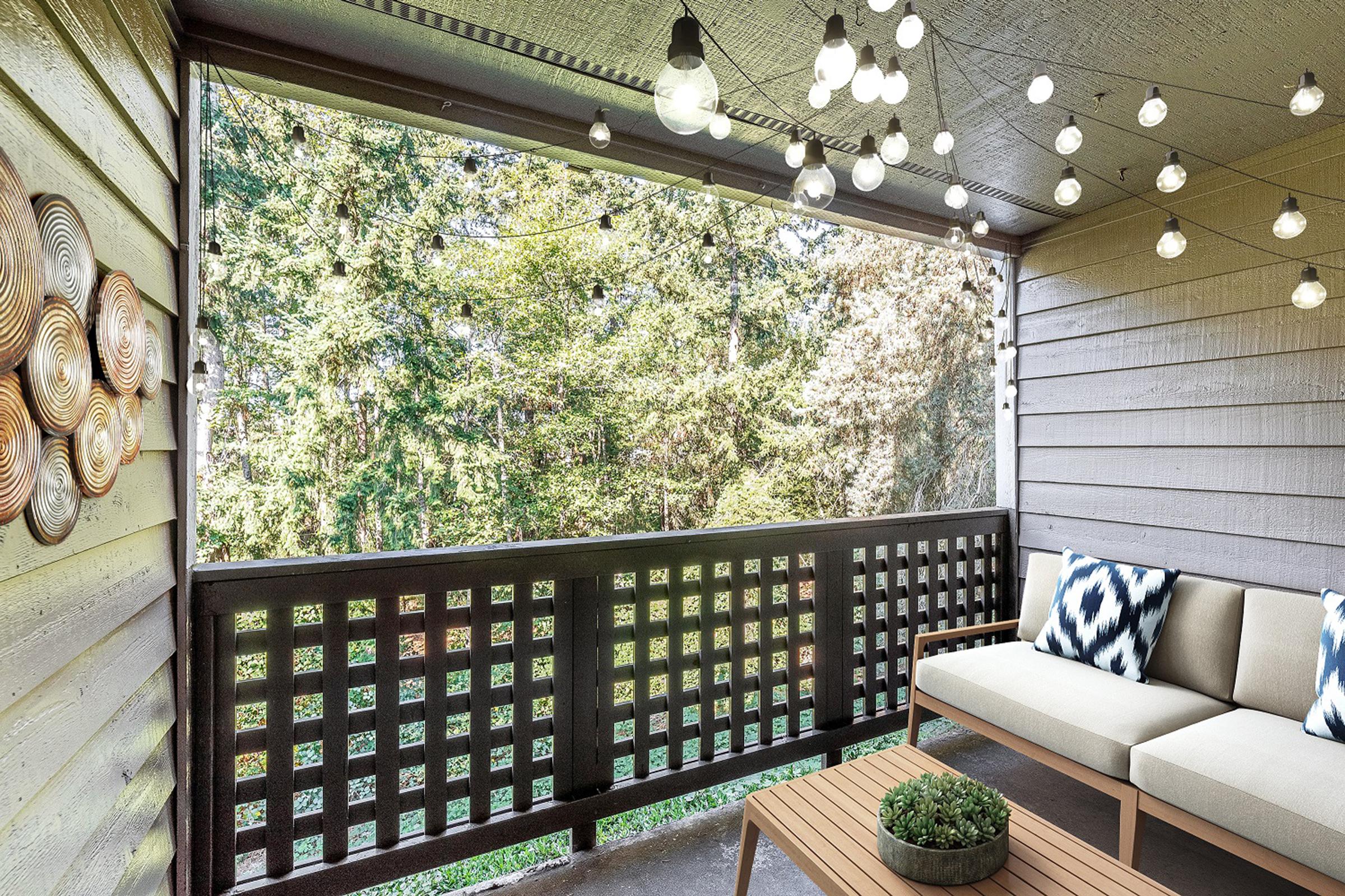 A cozy outdoor balcony with wooden paneling, featuring a comfortable beige sofa with patterned cushions and a small coffee table. The space is adorned with string lights, creating a warm ambiance, and overlooks greenery through a wooden railing. Decorative wall art adds an inviting touch.