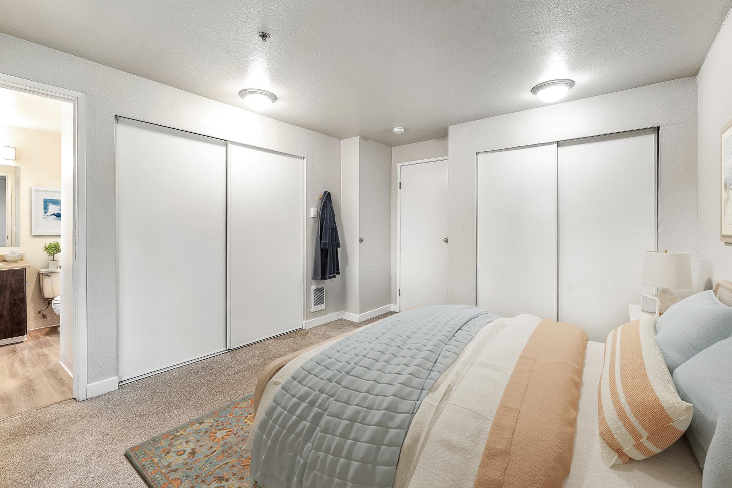 A cozy bedroom featuring a neatly made bed with light blue and beige bedding. The room has neutral-colored walls, two closet doors, and a decorative rug. A doorway leads to a bathroom area, and the space is illuminated by overhead lighting. The overall atmosphere is inviting and well-organized.
