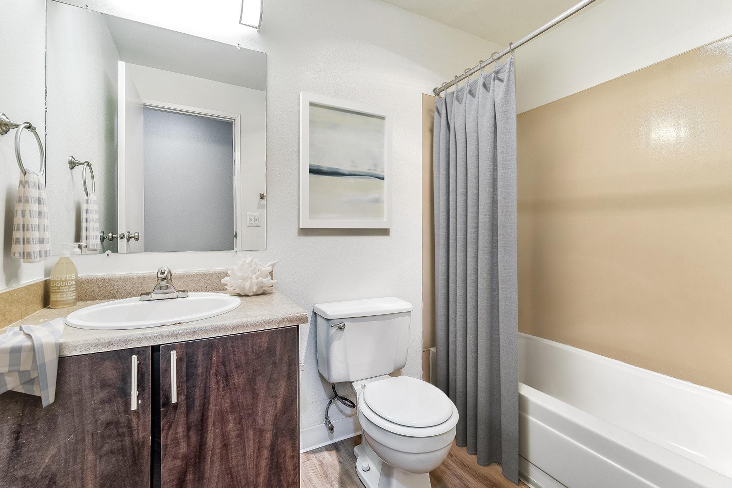 Modern bathroom featuring a white sink with a mirror above, a beige shower curtain, a bathtub, and a toilet. Light wood flooring adds warmth, with a simple piece of wall art and towels hanging nearby. The overall color scheme is neutral and clean, creating a serene atmosphere.