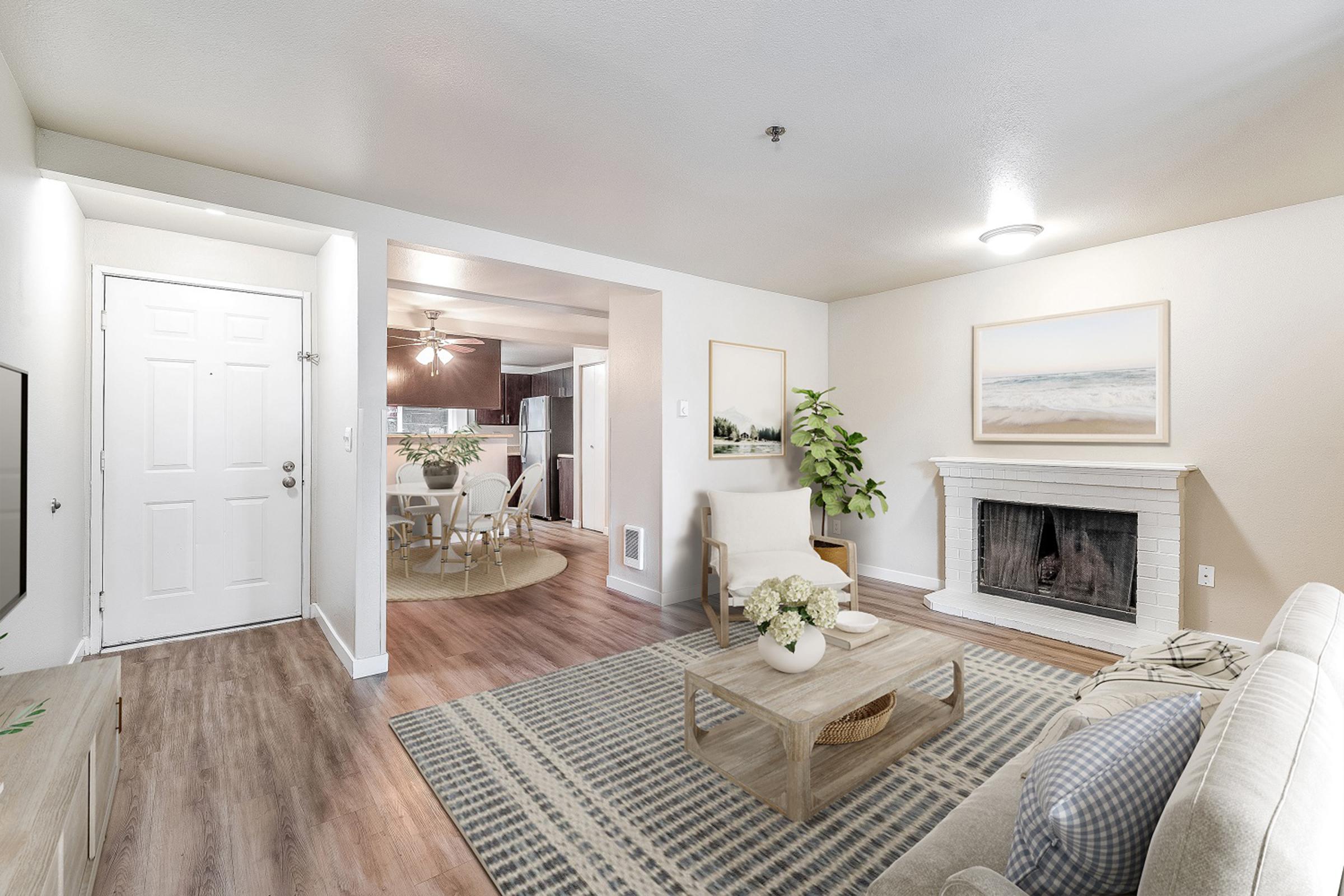 A bright and cozy living room featuring a light-colored couch, a coffee table on a patterned rug, and a decorative fireplace. The walls are painted in neutral tones, with a large piece of artwork above the fireplace. A doorway leads to a kitchen area, with a plant adding a touch of greenery to the space.
