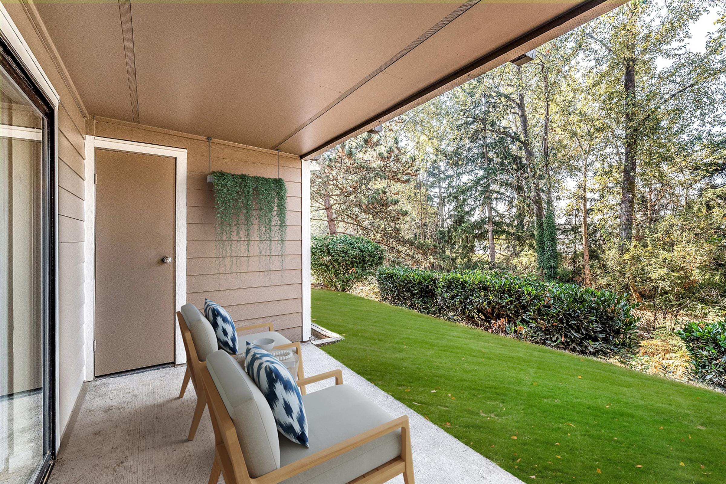 A covered patio featuring two light-colored chairs with decorative cushions, overlooking a lush green lawn and a densely wooded area beyond. The walls are a soft beige, and a small door is visible, along with hanging greenery. Sunlight filters through the trees, creating a serene outdoor atmosphere.
