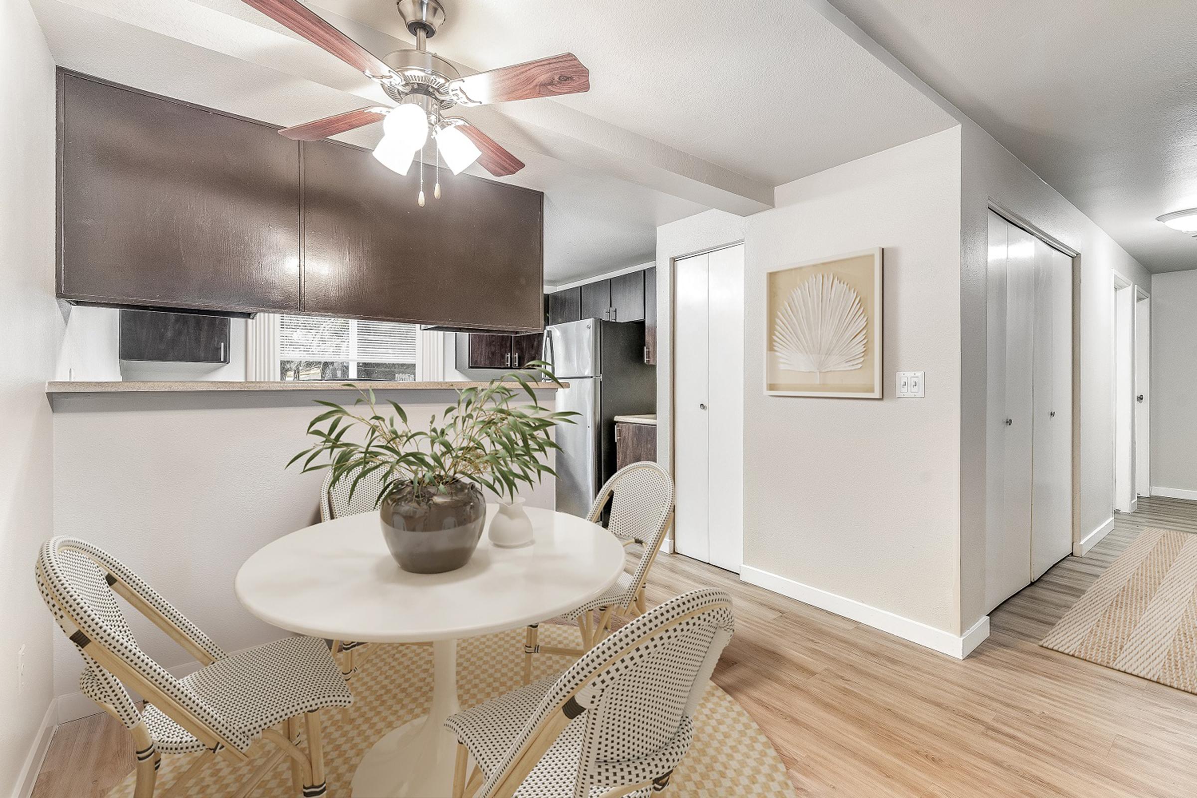 A cozy dining area featuring a round white table with four rattan chairs. A potted plant sits on the table. The background includes a partially visible kitchen with modern appliances and a hallway with sliding doors. Natural light filters in through a window covered by dark blinds.
