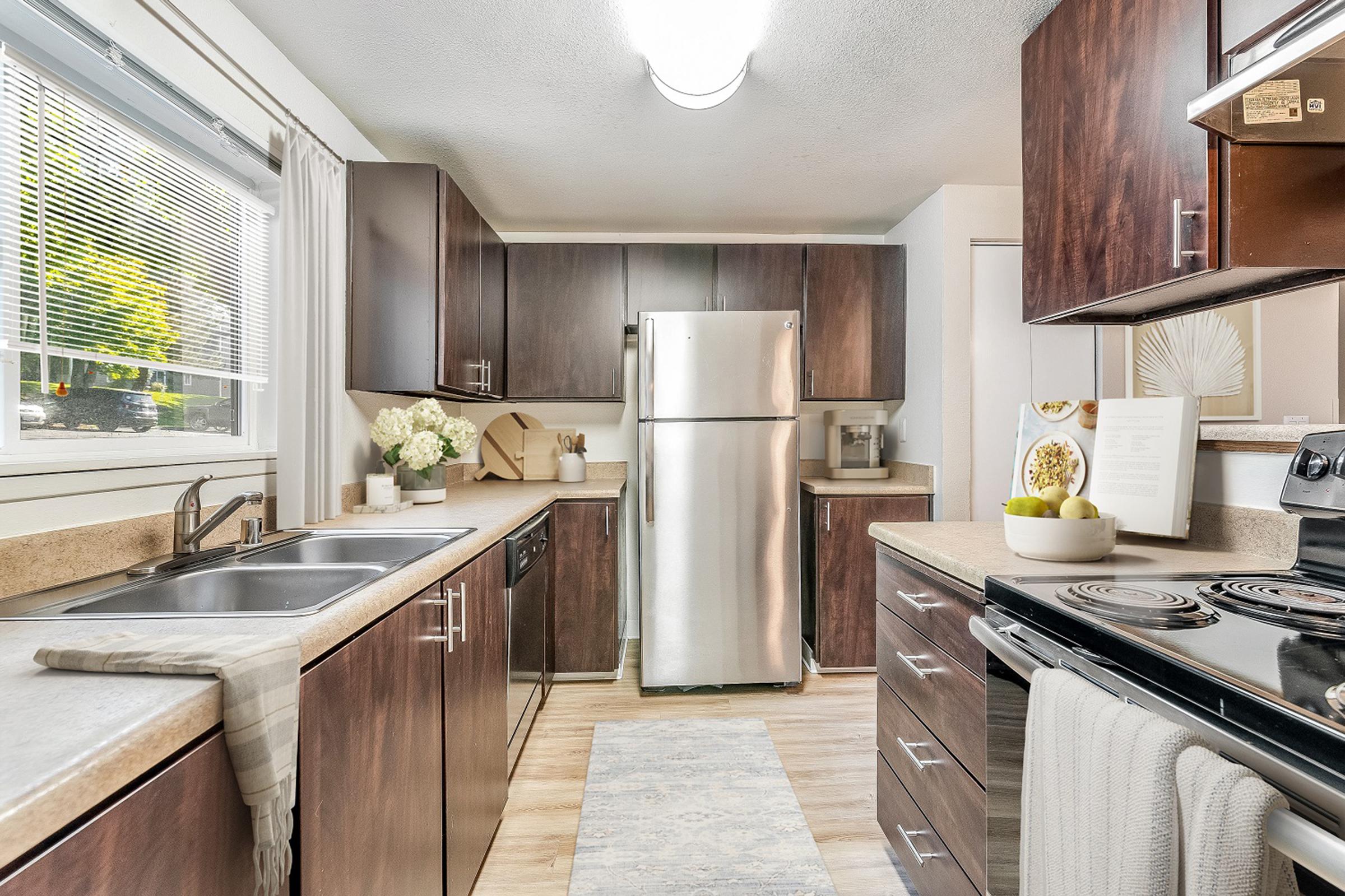 A modern kitchen featuring dark wood cabinets, a stainless steel refrigerator, and an oven. The countertop is made of a light material with a bowl of fruit and decorative items. Natural light streams in through a window, illuminating the space with a cozy and inviting atmosphere.