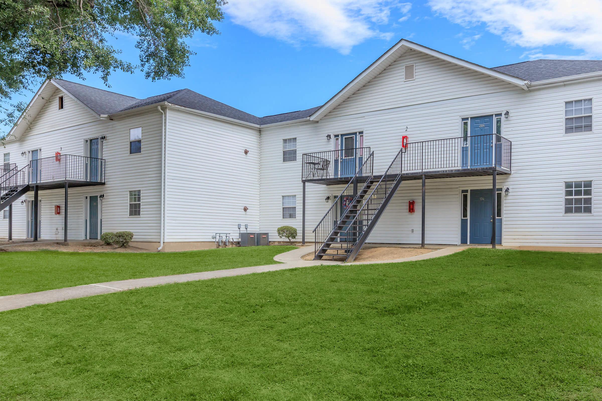 Two-story residential building with multiple units, featuring white siding and blue doors. Each unit has a balcony, and the landscape includes a green lawn and a path leading to the entrance. There are outdoor fire extinguishers visible on the wall. The sky is partly cloudy.