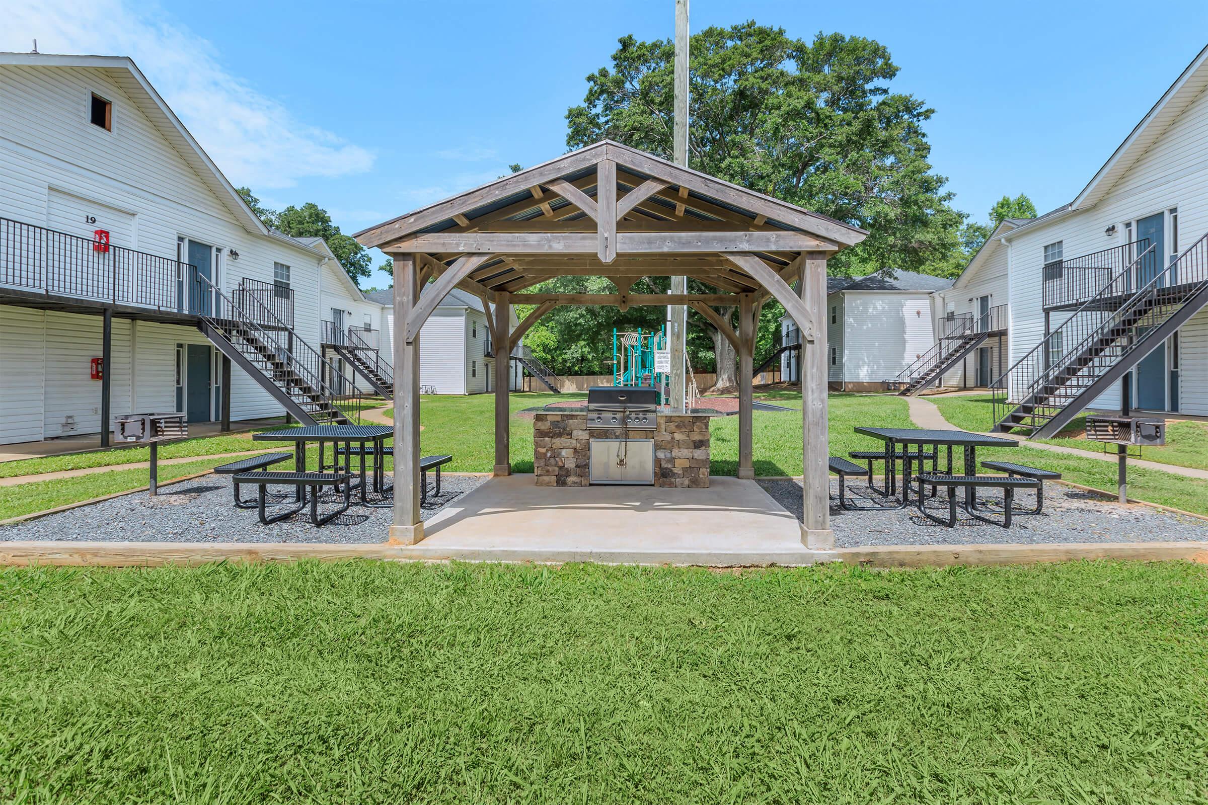 A spacious outdoor gathering area featuring a wooden pavilion with a stone grill, surrounded by picnic tables, set in a grassy courtyard. In the background, there are two-story apartment buildings with balconies and a playground area visible nearby under a clear blue sky.