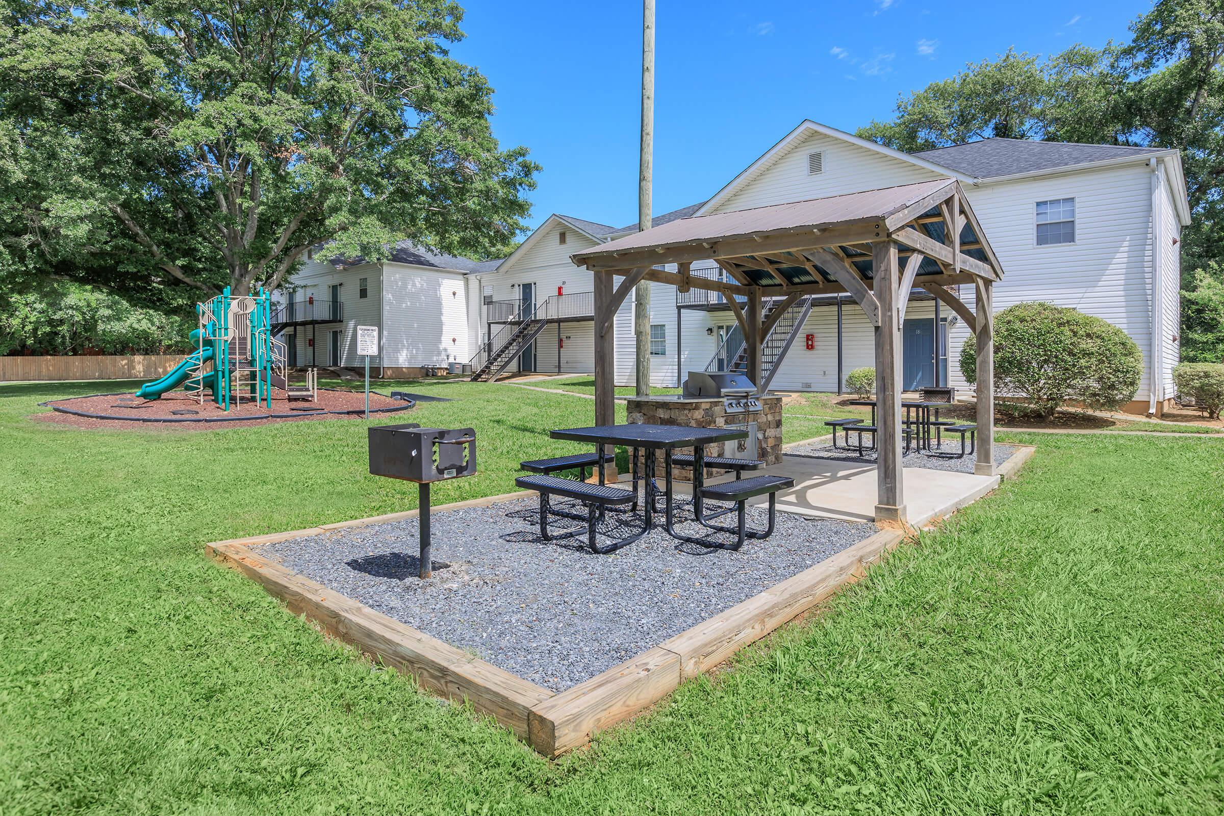 A picnic area with a wooden gazebo featuring picnic tables and a grill, surrounded by green grass. In the background, there are two white apartment buildings and a playground with slides and climbing structures under a blue sky.